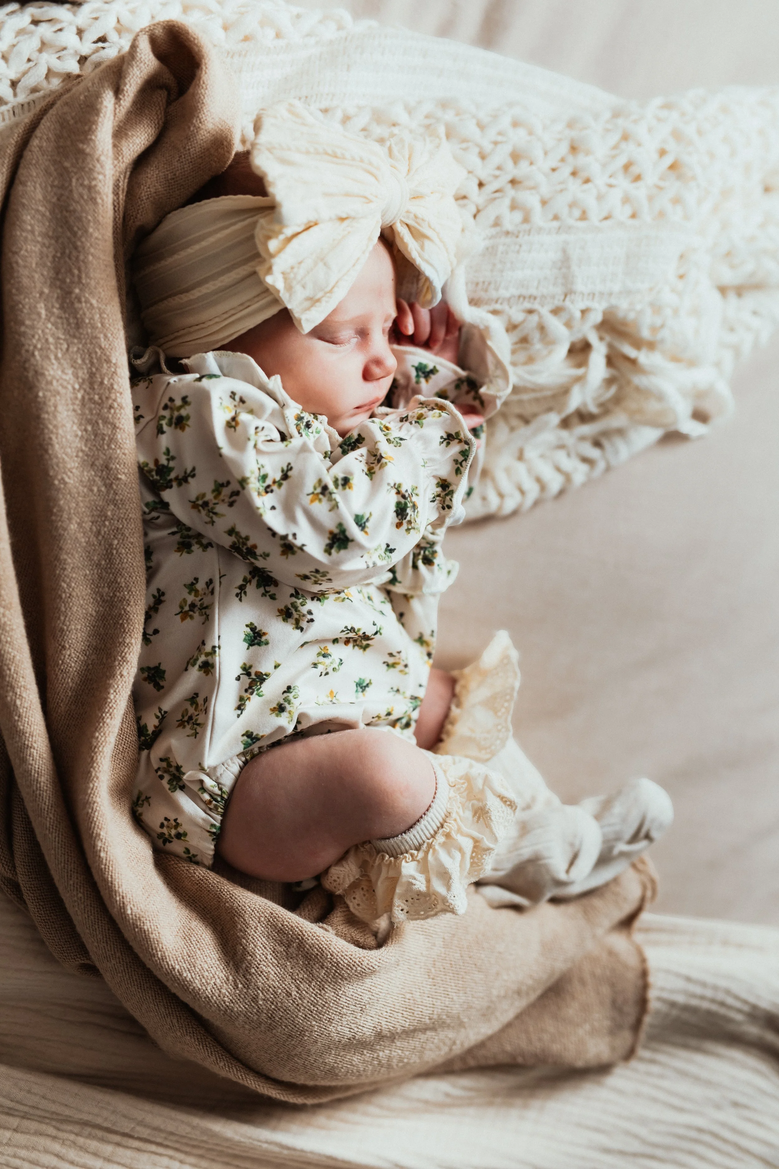 A sleeping baby girl wearing a floral pajama, beanie, and a large bow headband, nestled in a soft, cream-colored blanket on a beige cushion.