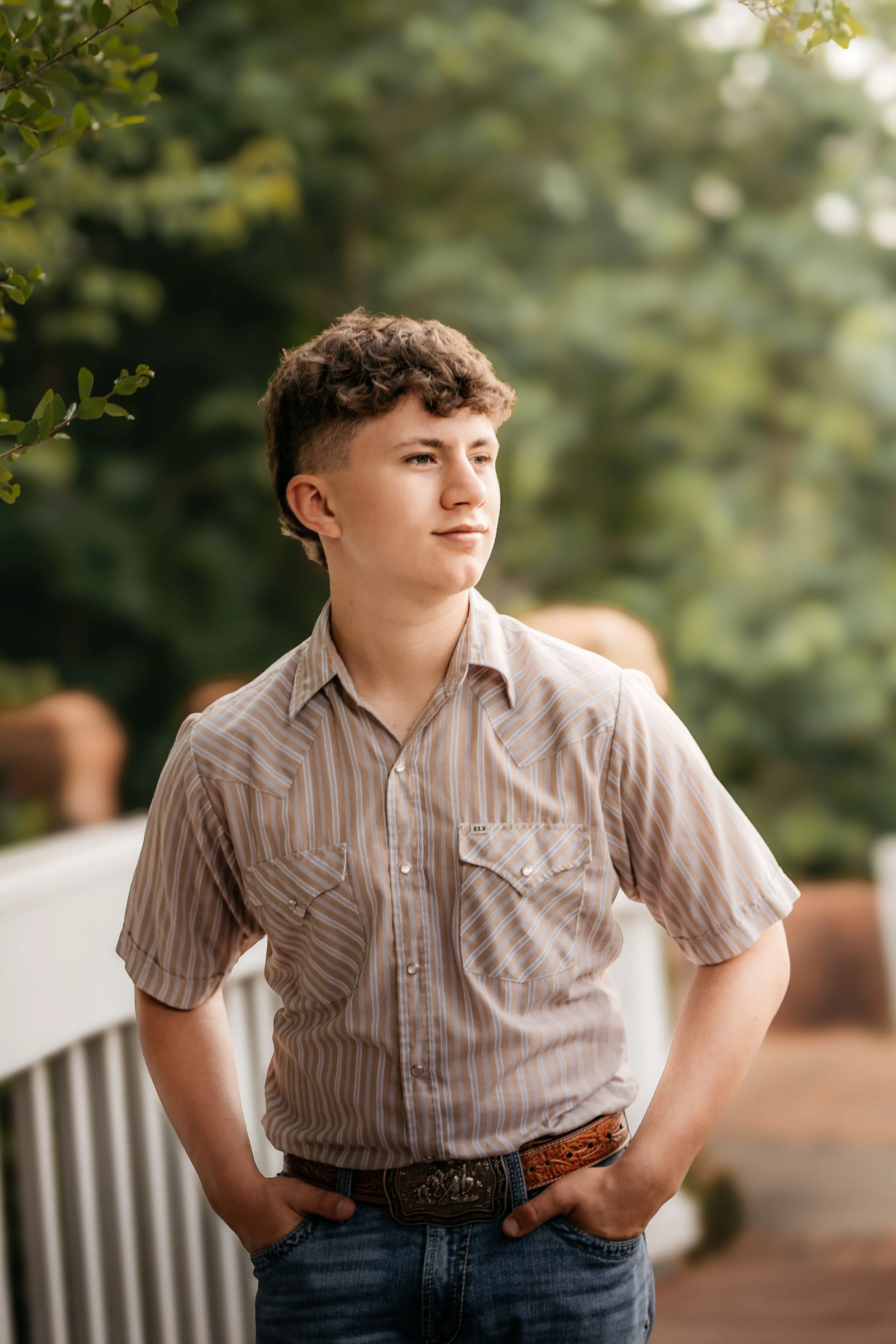 A young man with curly brown hair wearing a beige and brown striped short-sleeve button-up shirt, blue jeans, a belt with a decorative buckle, standing outdoors near a white fence, with green trees and blurred background.