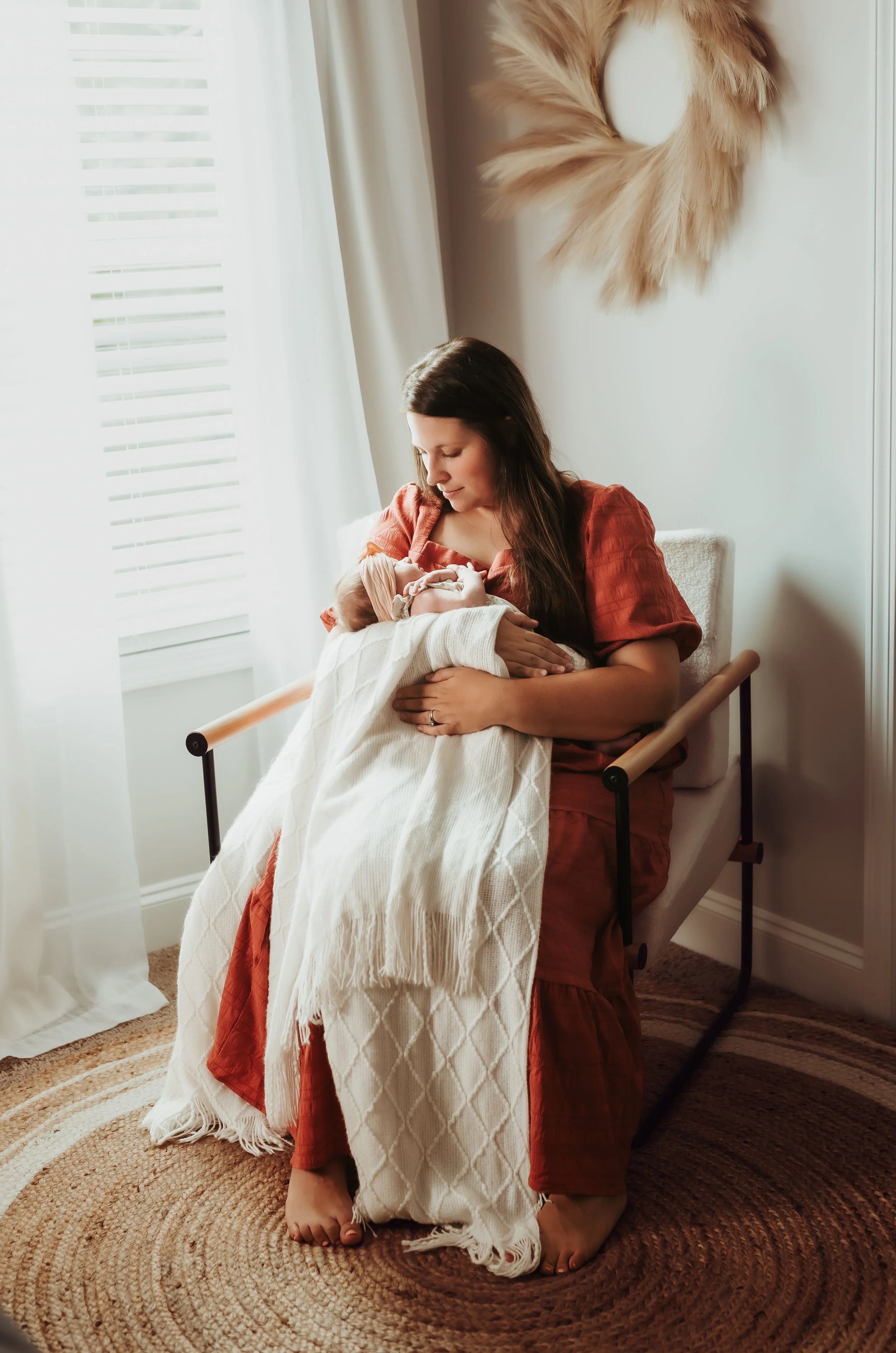 A woman sitting on a bed nursing a newborn baby wrapped in a cream-colored blanket. The woman is wearing a reddish-brown dress and has long dark hair. The room has white walls, a wreath with faux fur hanging on the wall, and a window with white curta