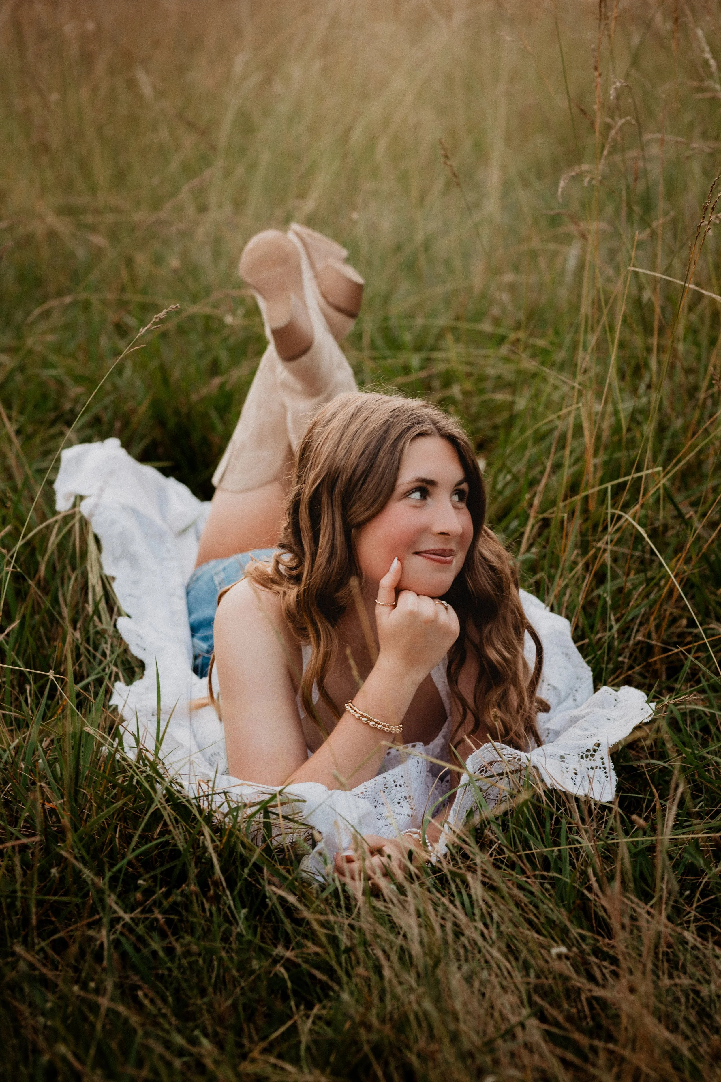 A young woman with long wavy brown hair lying in tall grass, wearing a white lace dress and beige cowboy boots, looking thoughtfully to the side.