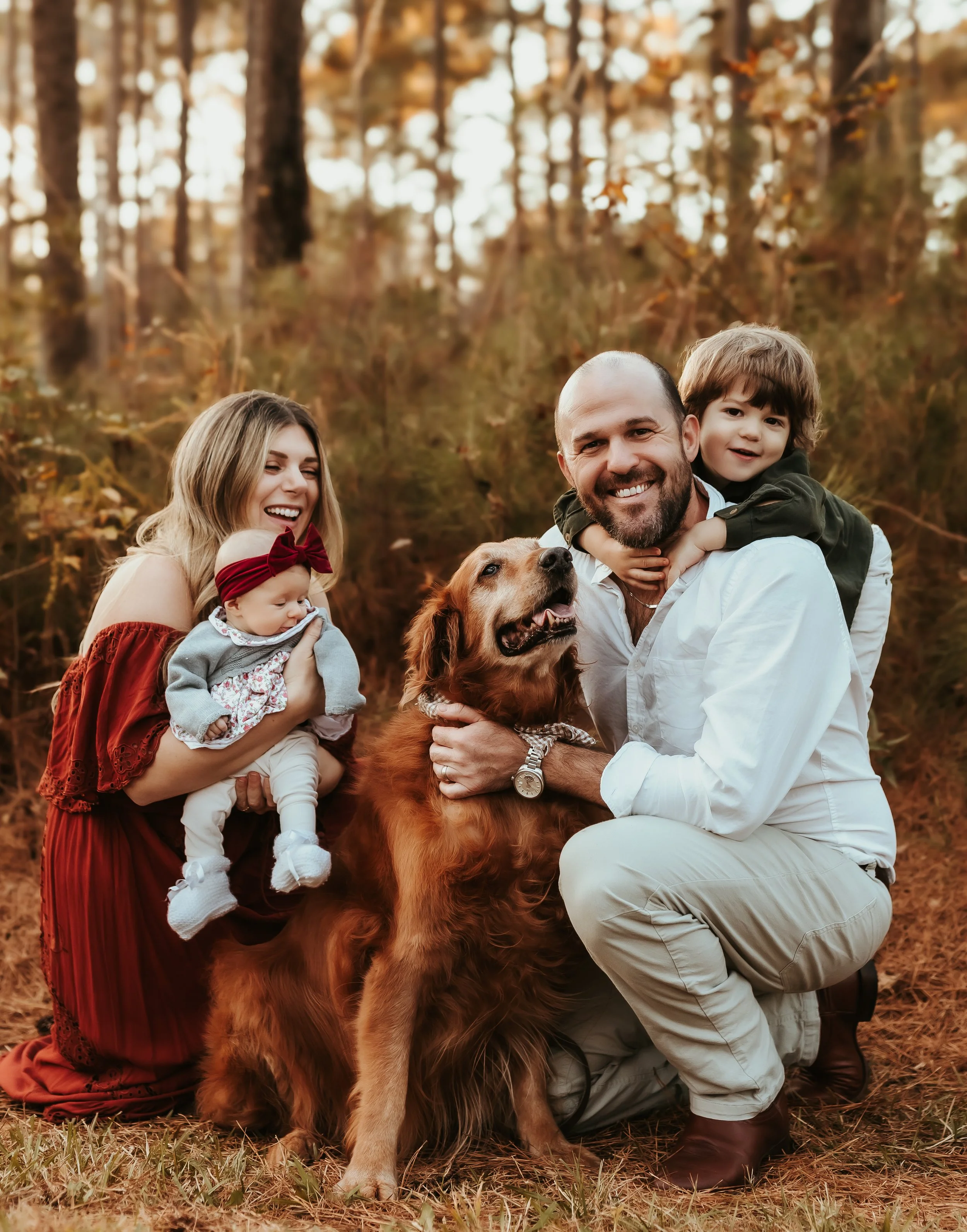 Family of four with a dog in a wooded park during fall, smiling and enjoying the outdoors.