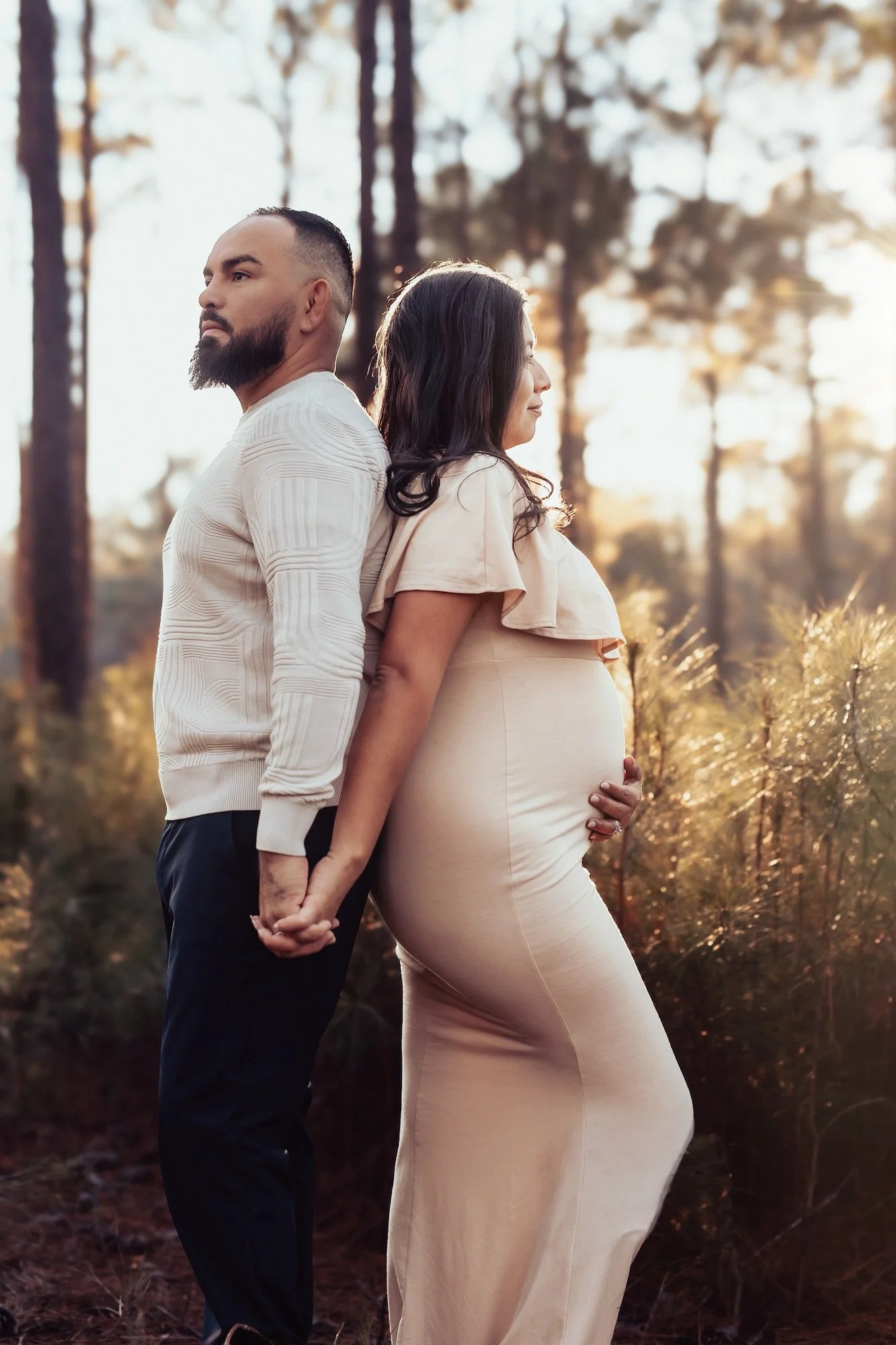 Pregnant woman and her partner holding hands with their backs against each other in a forest during sunset.