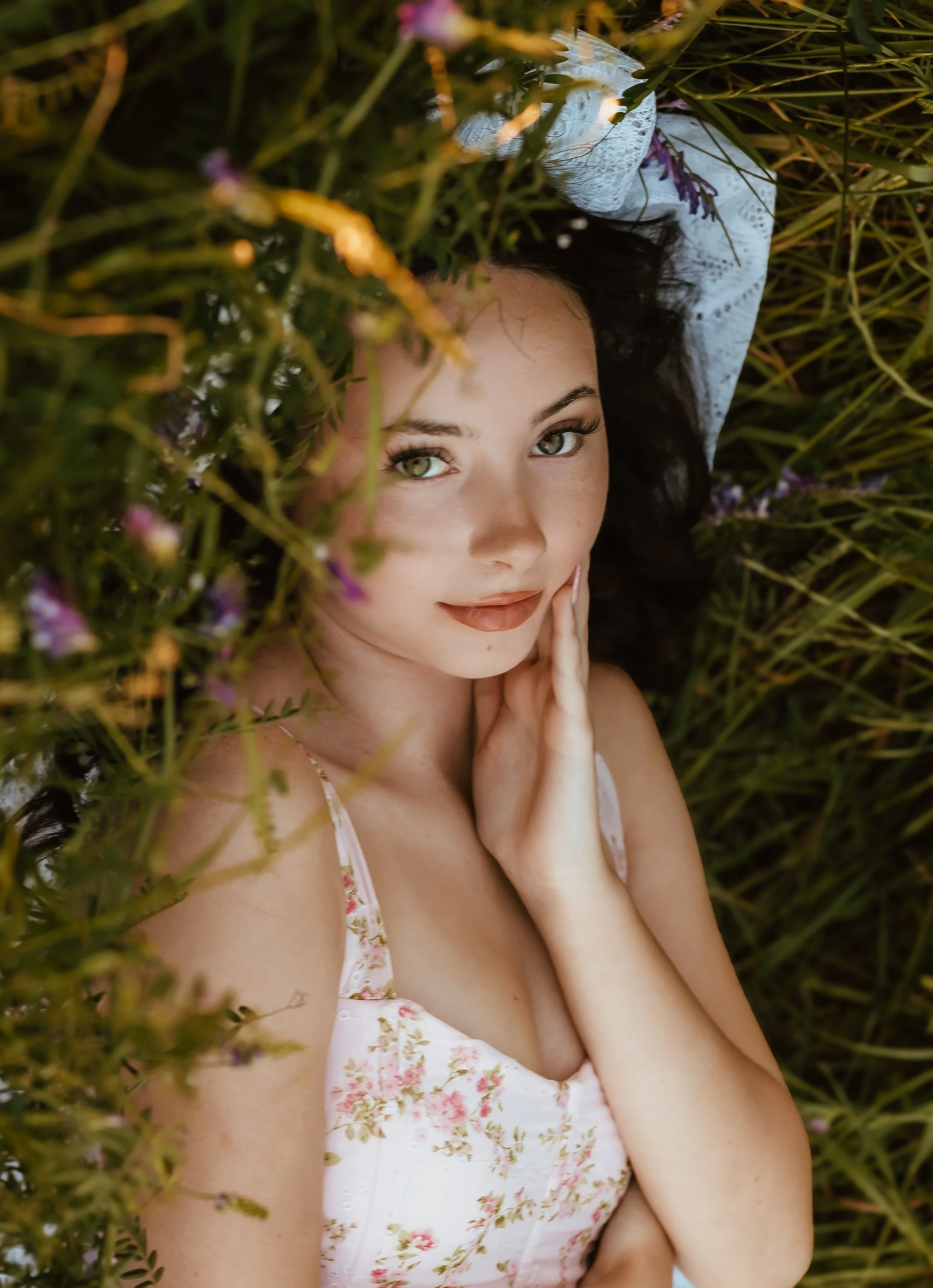 A young woman with black hair and light skin lying among green plants and purple flowers, looking at the camera with her hand gently touching her face.