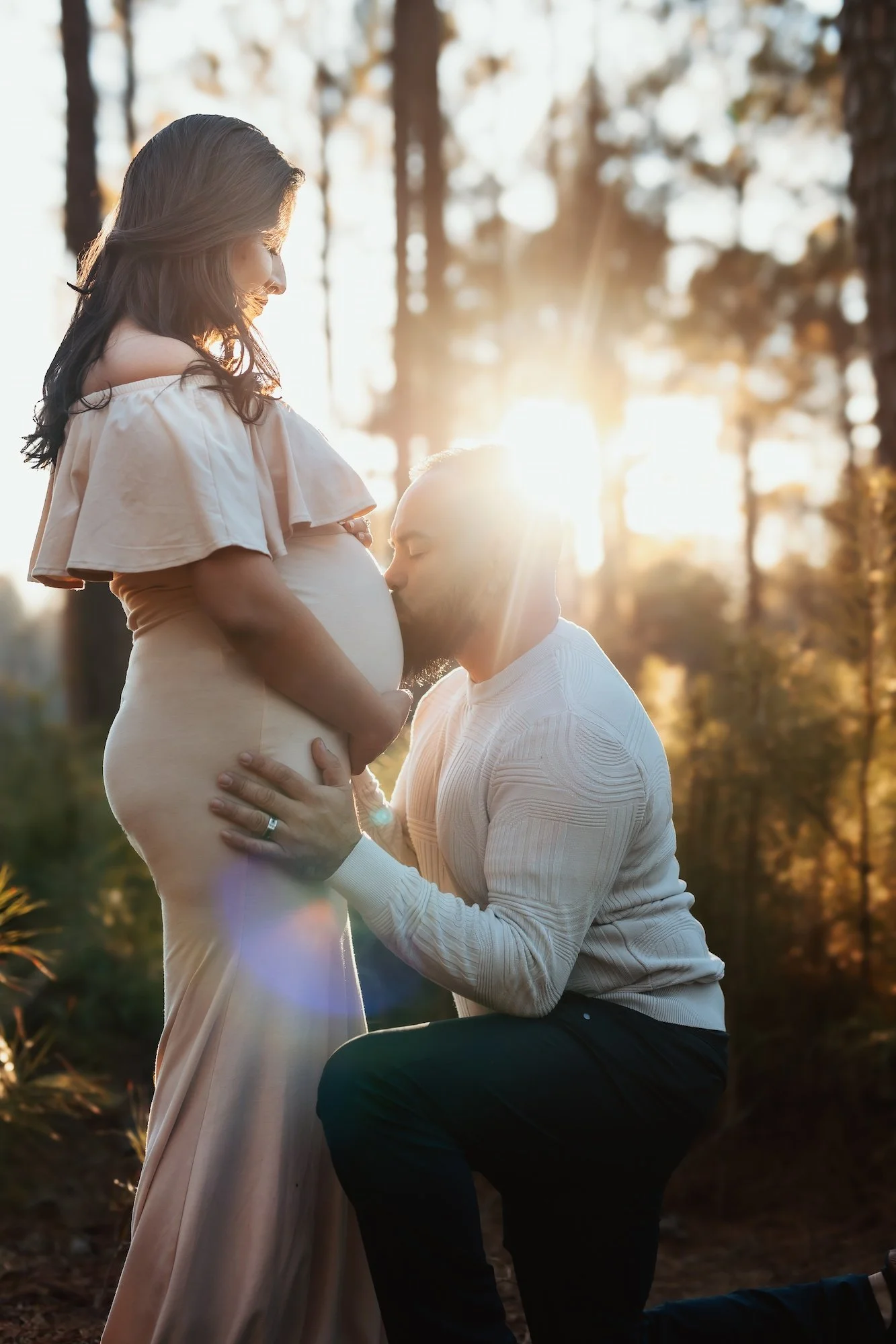A pregnant woman standing outdoors in a forest at sunset, with a man kneeling and kissing her belly.
