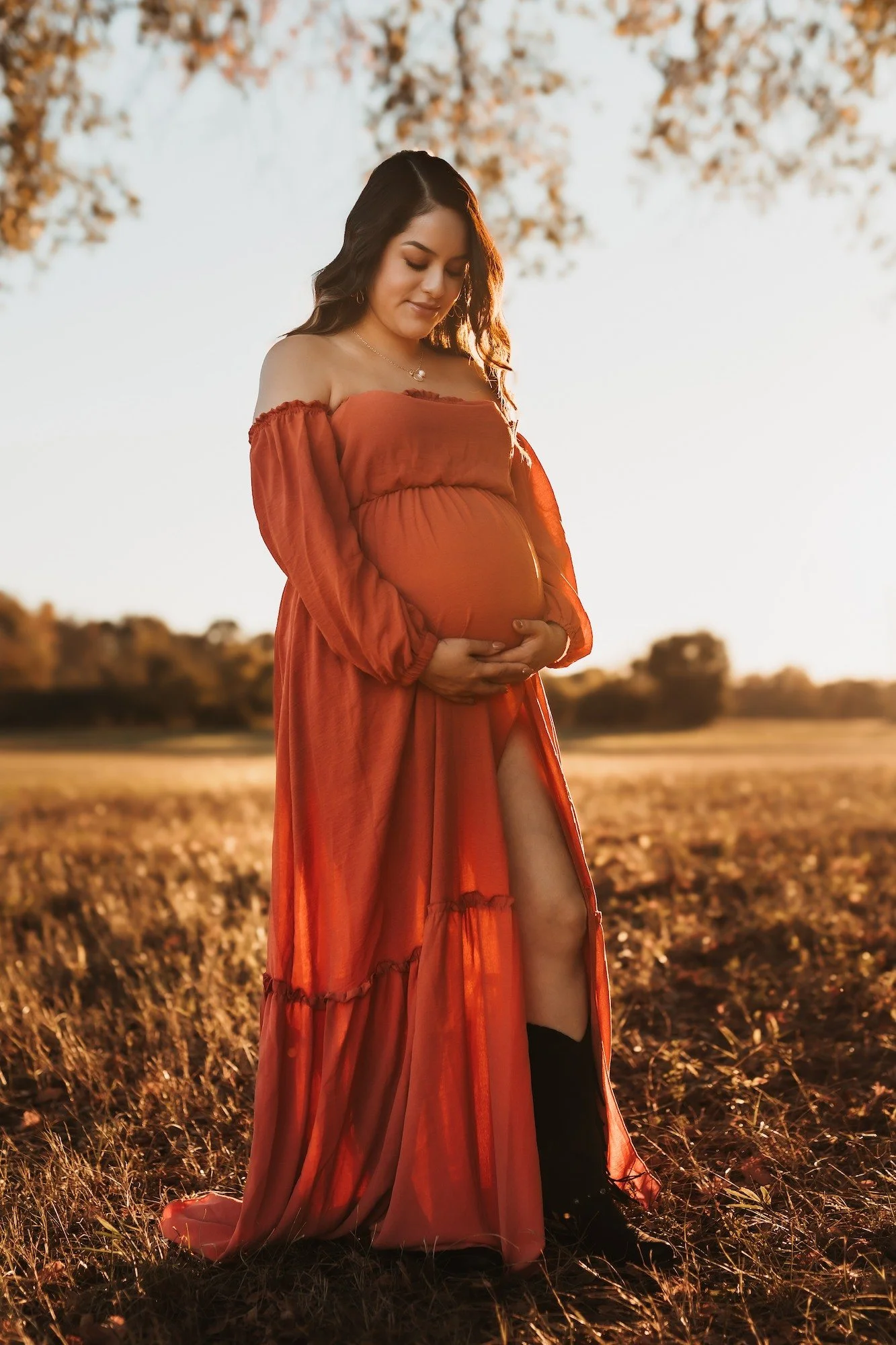 A pregnant woman in a flowing red dress stands outdoors in a field during sunset, gently holding her belly and smiling softly.
