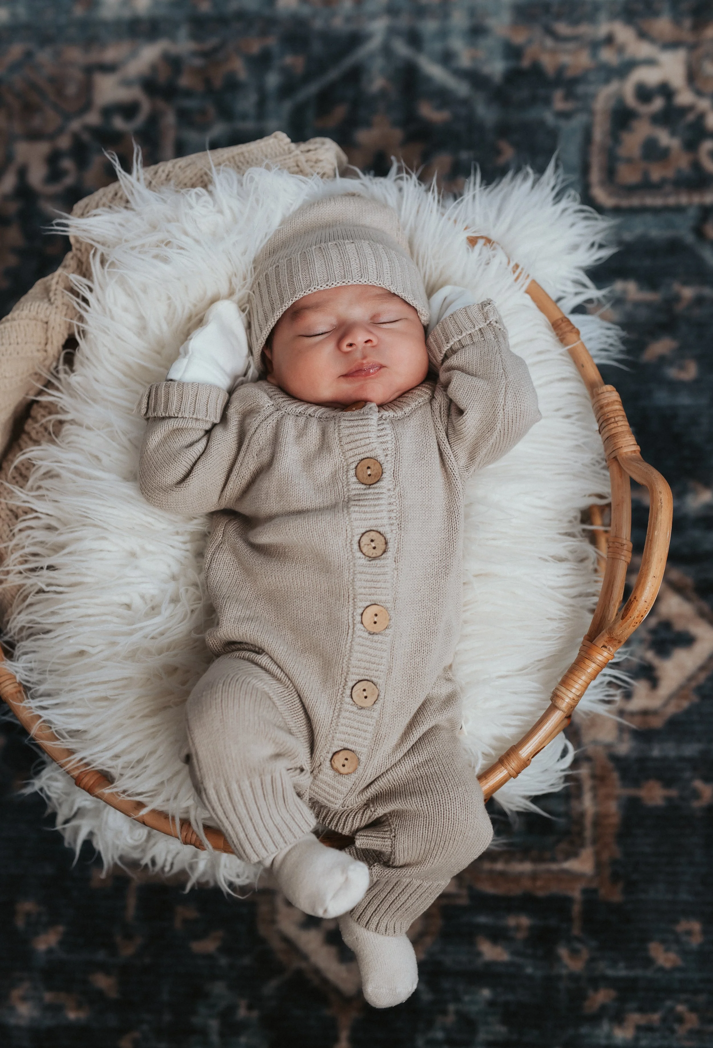 A sleeping baby in a beige knitted onesie with buttons, wearing a beige beanie, lying on a fluffy white rug in a wicker basket, on a dark patterned rug.