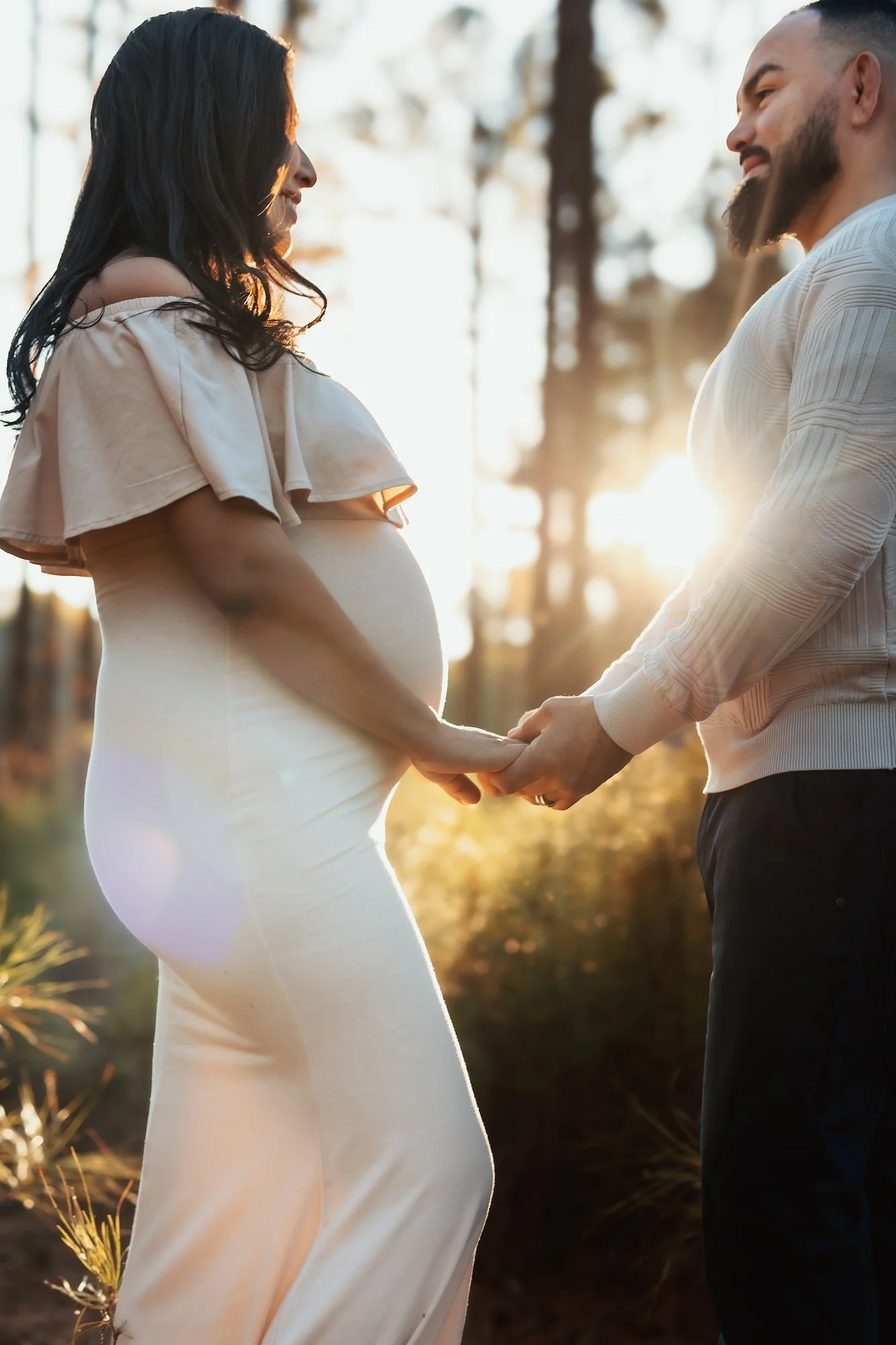 Pregnant woman and man holding hands outdoors at sunset, facing each other, with trees in the background.