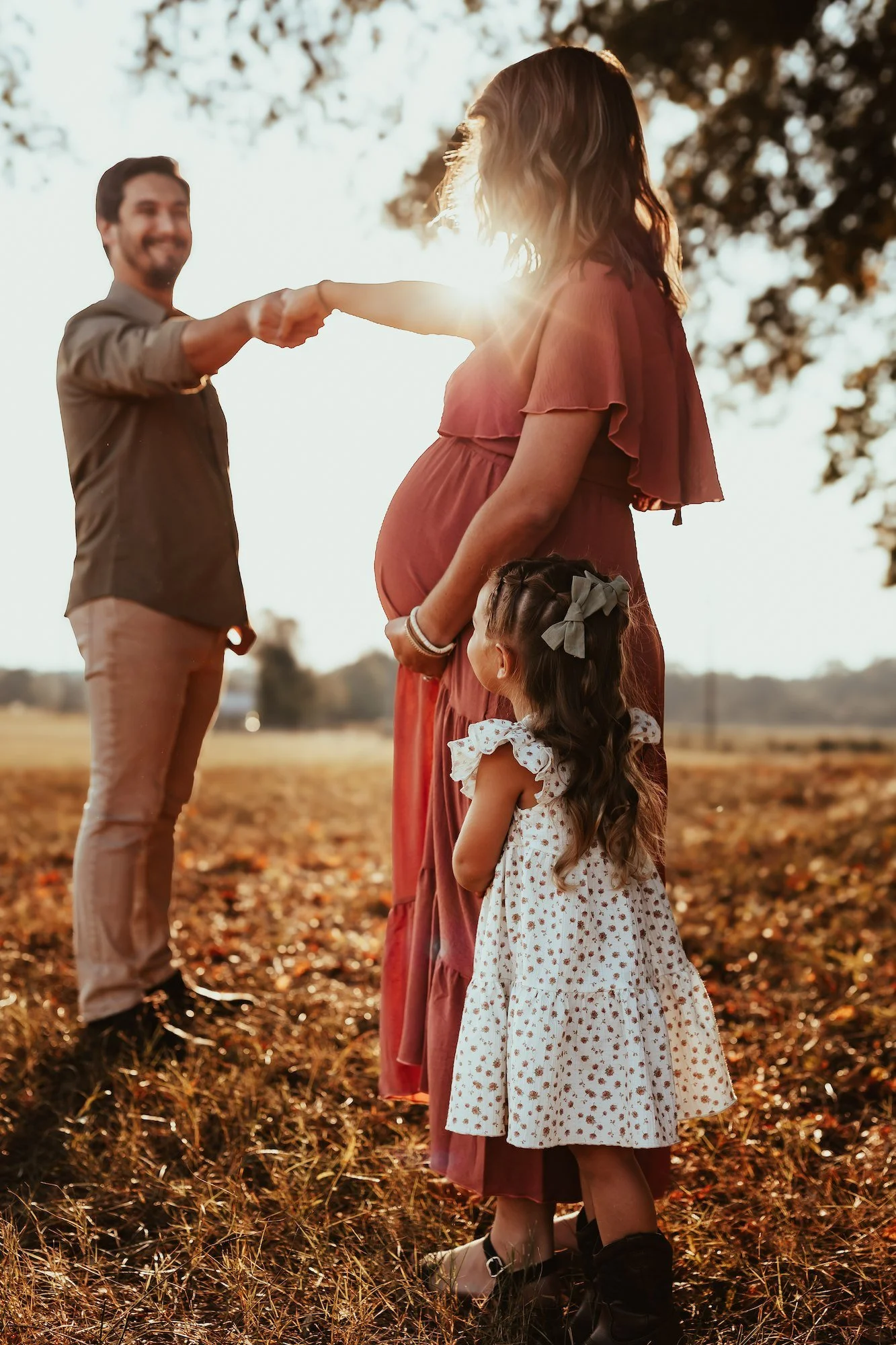A pregnant woman standing outdoors in a field with sunlight shining behind her, holding her belly. A man is holding one of her outstretched hands, while a young girl in a white dress with polka dots stands beside her, looking up at the woman. The sce