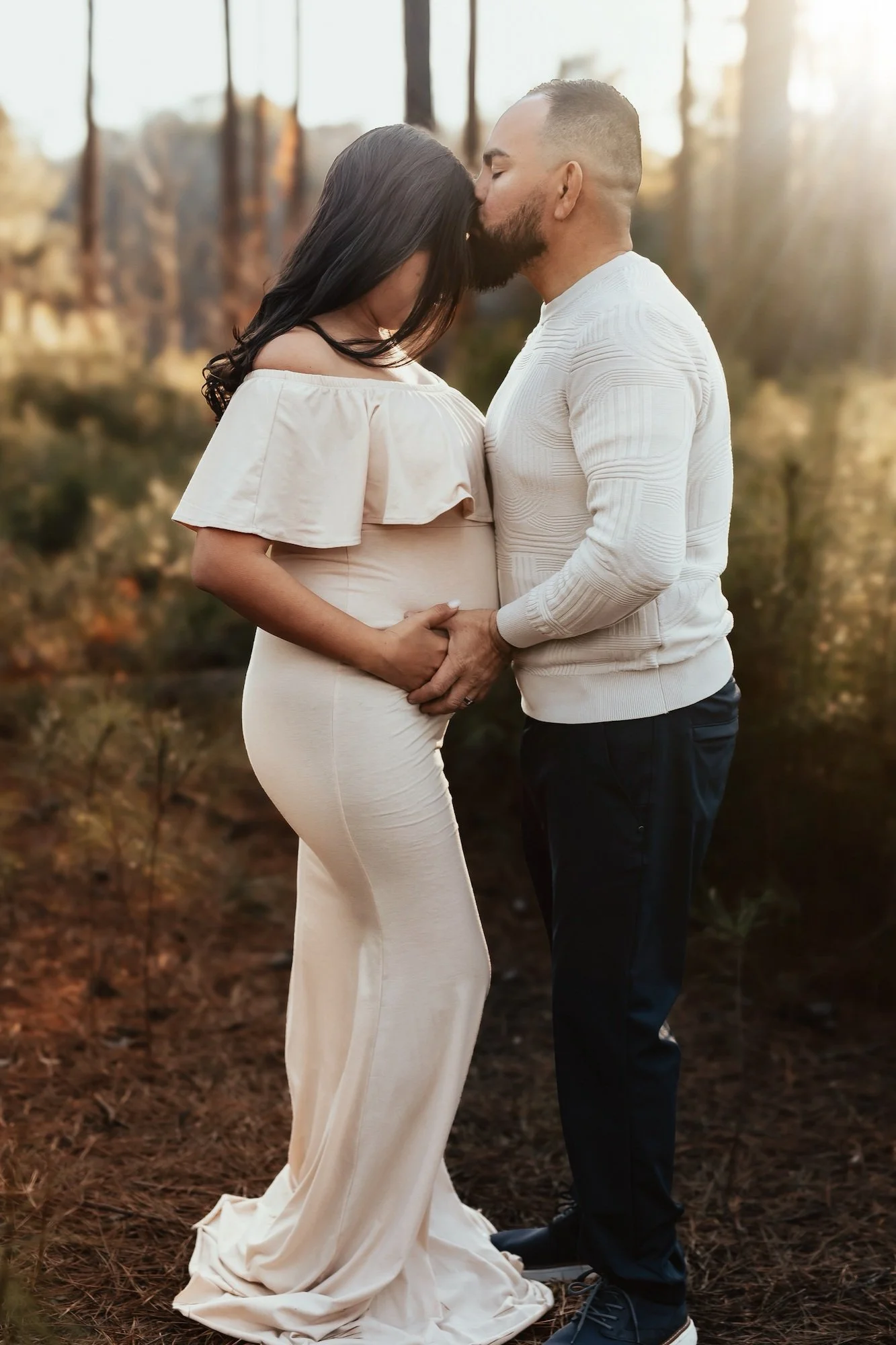 A couple is kissing and holding hands outdoors during sunset. The woman wears a cream-colored gown with ruffled sleeves, and the man wears a white sweater and dark pants. They stand on a forest floor with blurred trees in the background.
