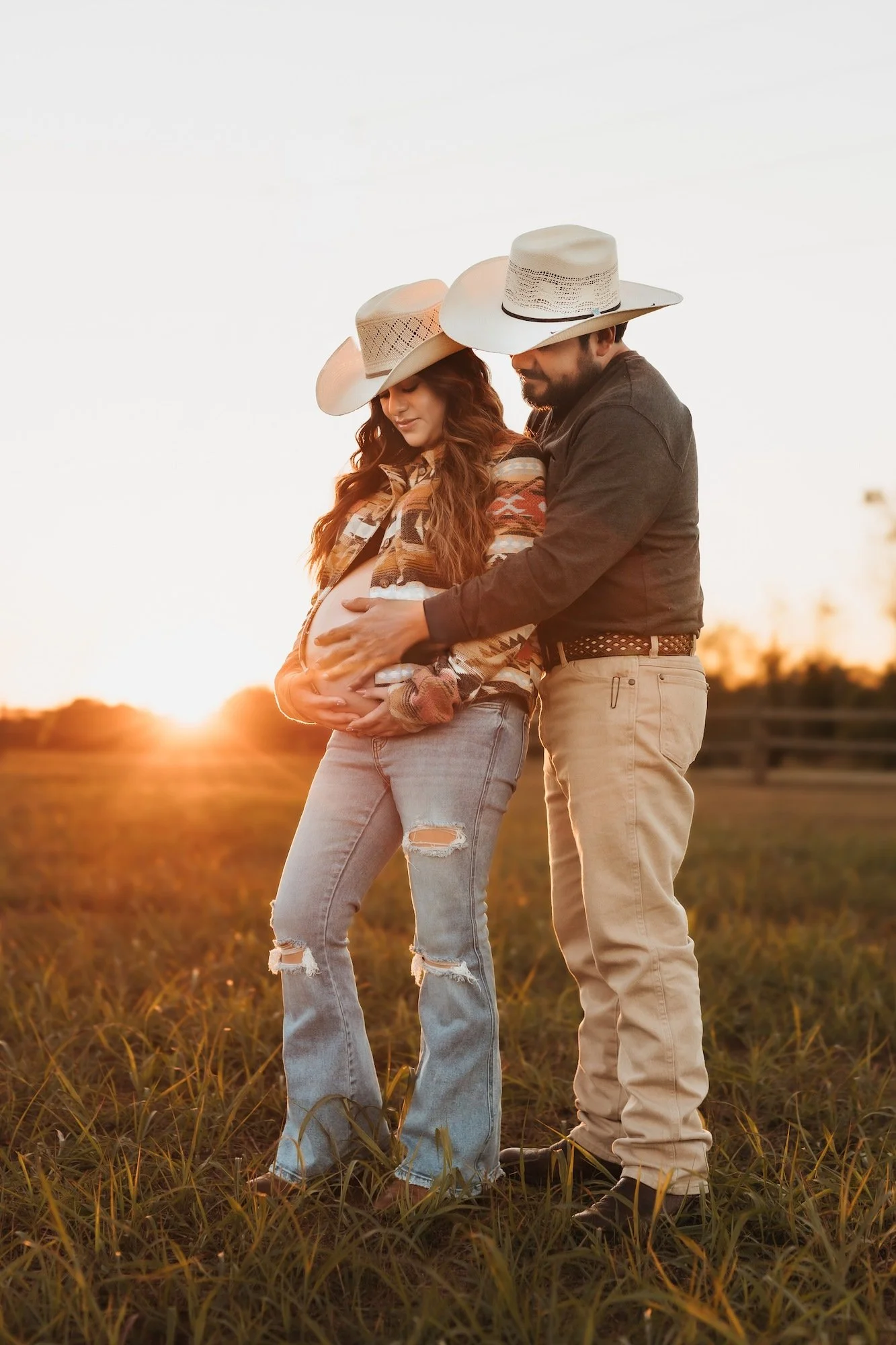 A pregnant woman and a man, both wearing cowboy hats, standing together in a field at sunset. The man is touching the woman's belly, and they are both looking down at it with affection.