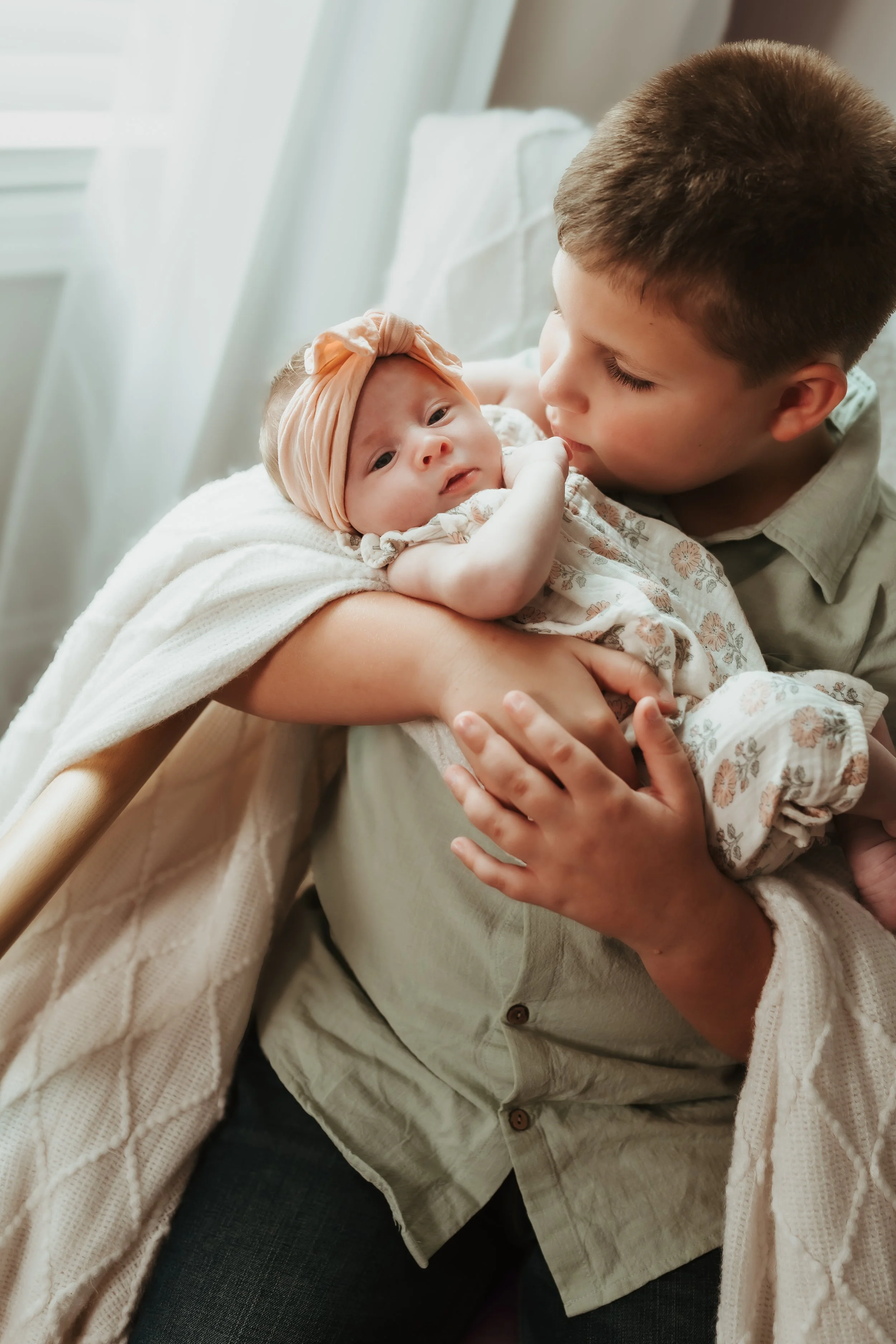 A young boy holding a newborn baby girl in his arms. The baby is wearing a peach-colored headband and a floral outfit. They are indoors near a window with white curtains.