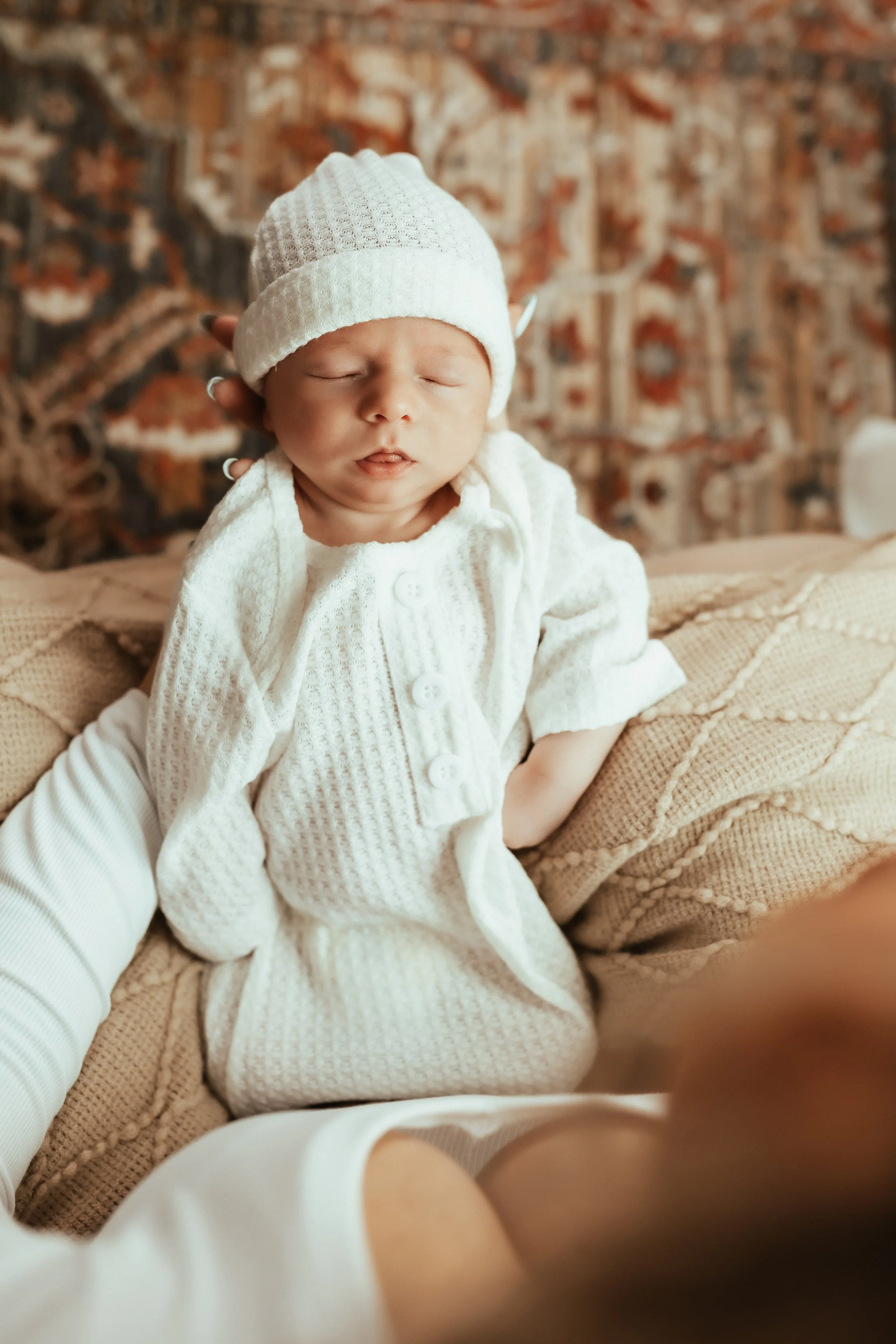 Close-up of a sleeping newborn baby wearing a white knit hat and white sweater, with an abstract patterned background.