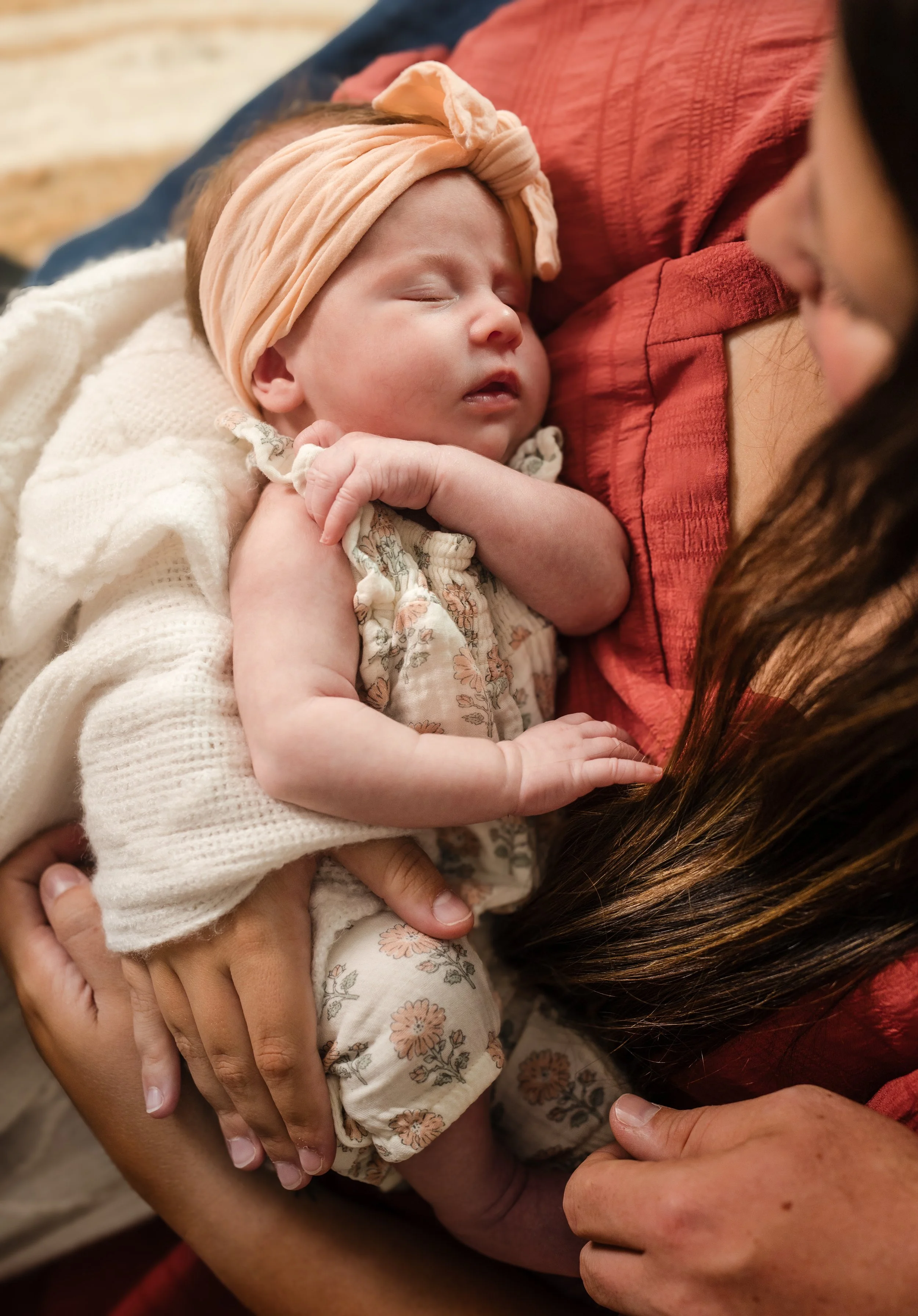 Close-up of a sleeping baby with a peach headband, cradled in a person's arms, with another person gently touching the baby's hand.