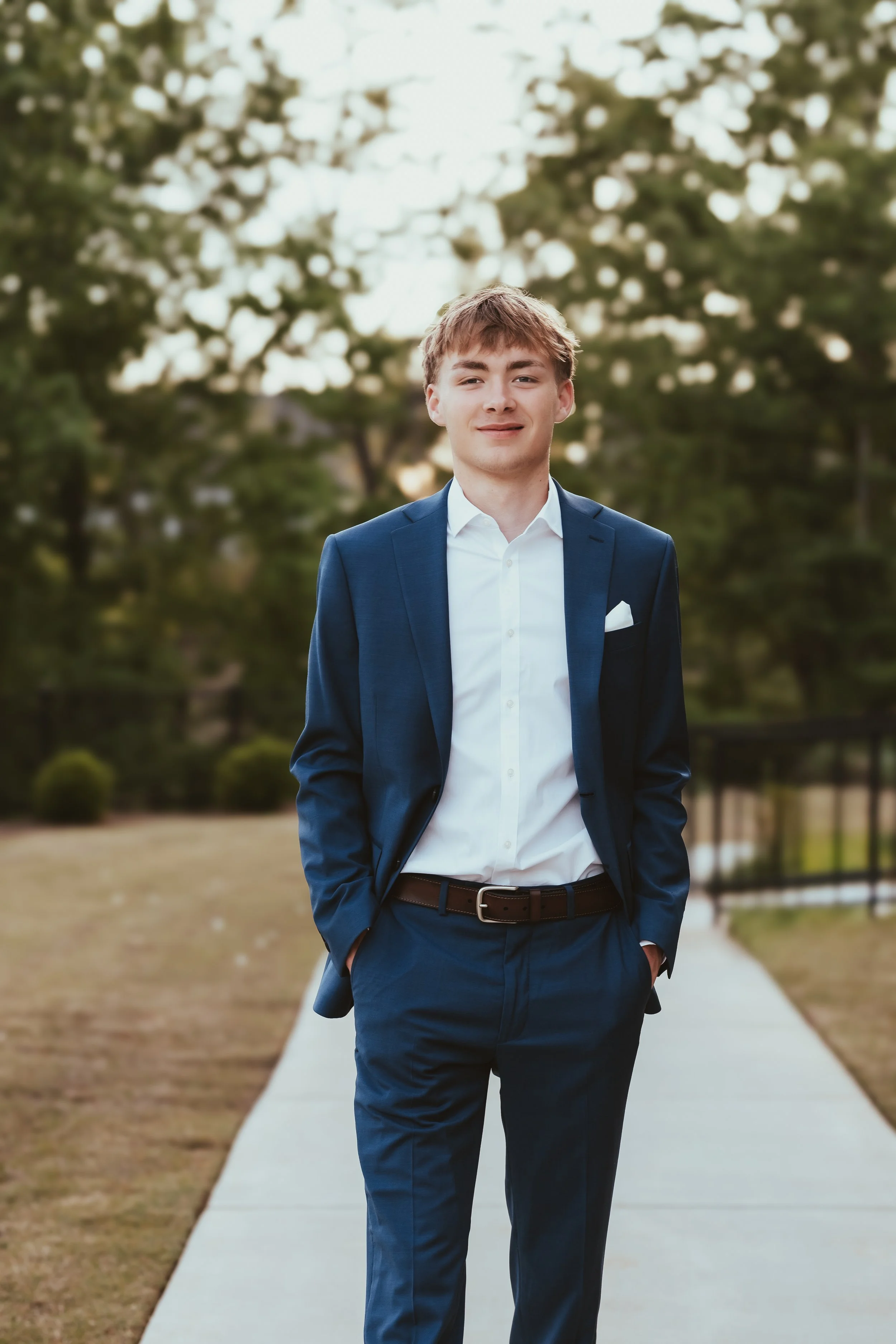 Young man in a blue suit with a white shirt and pocket square, standing outdoors on a park sidewalk during daytime, with trees in the background.