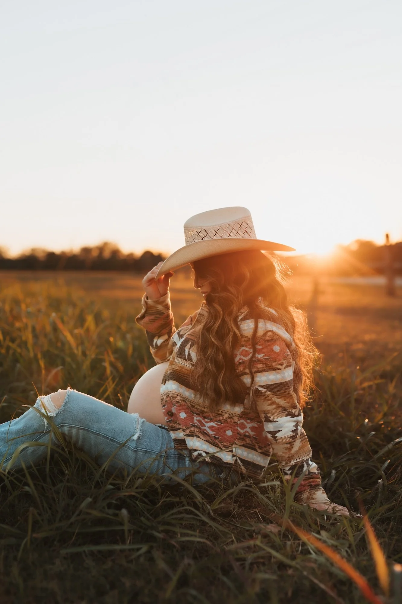 A pregnant woman sitting in a field at sunset, wearing a cowboy hat and patterned sweater.