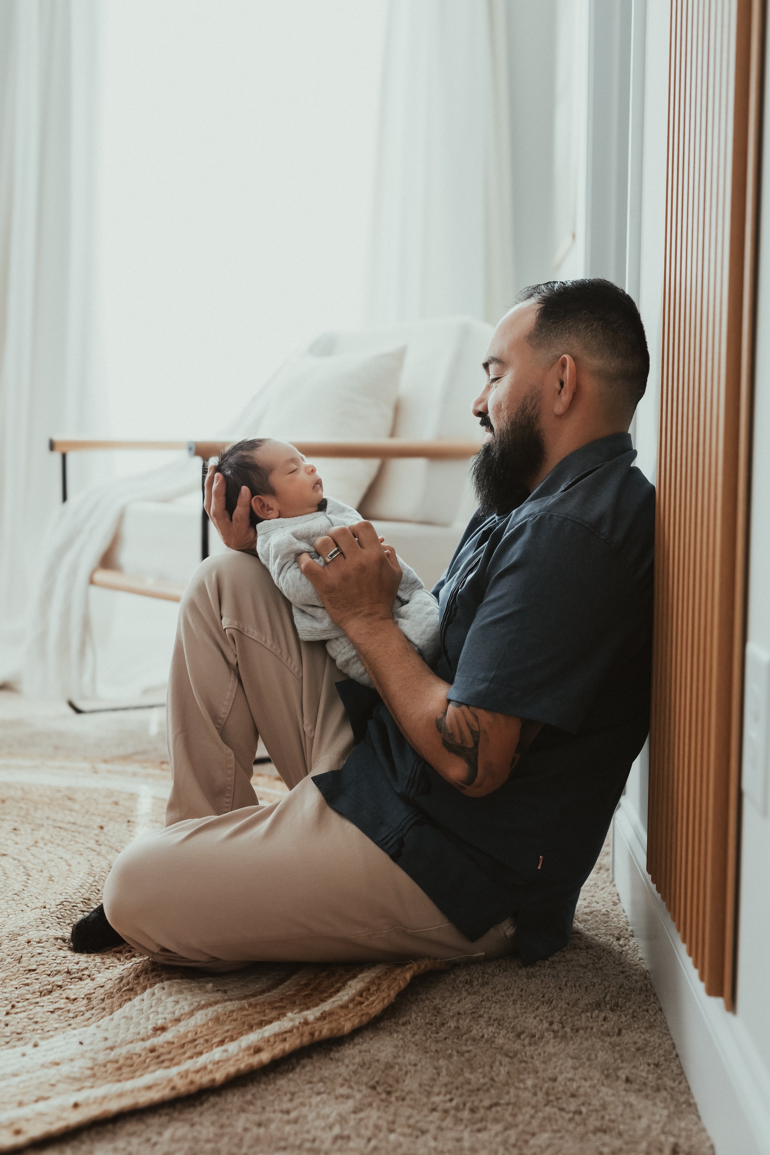A man kneeling on a rug, holding a sleeping baby in his hands and looking at the baby affectionately in a cozy room with a bed and curtains.
