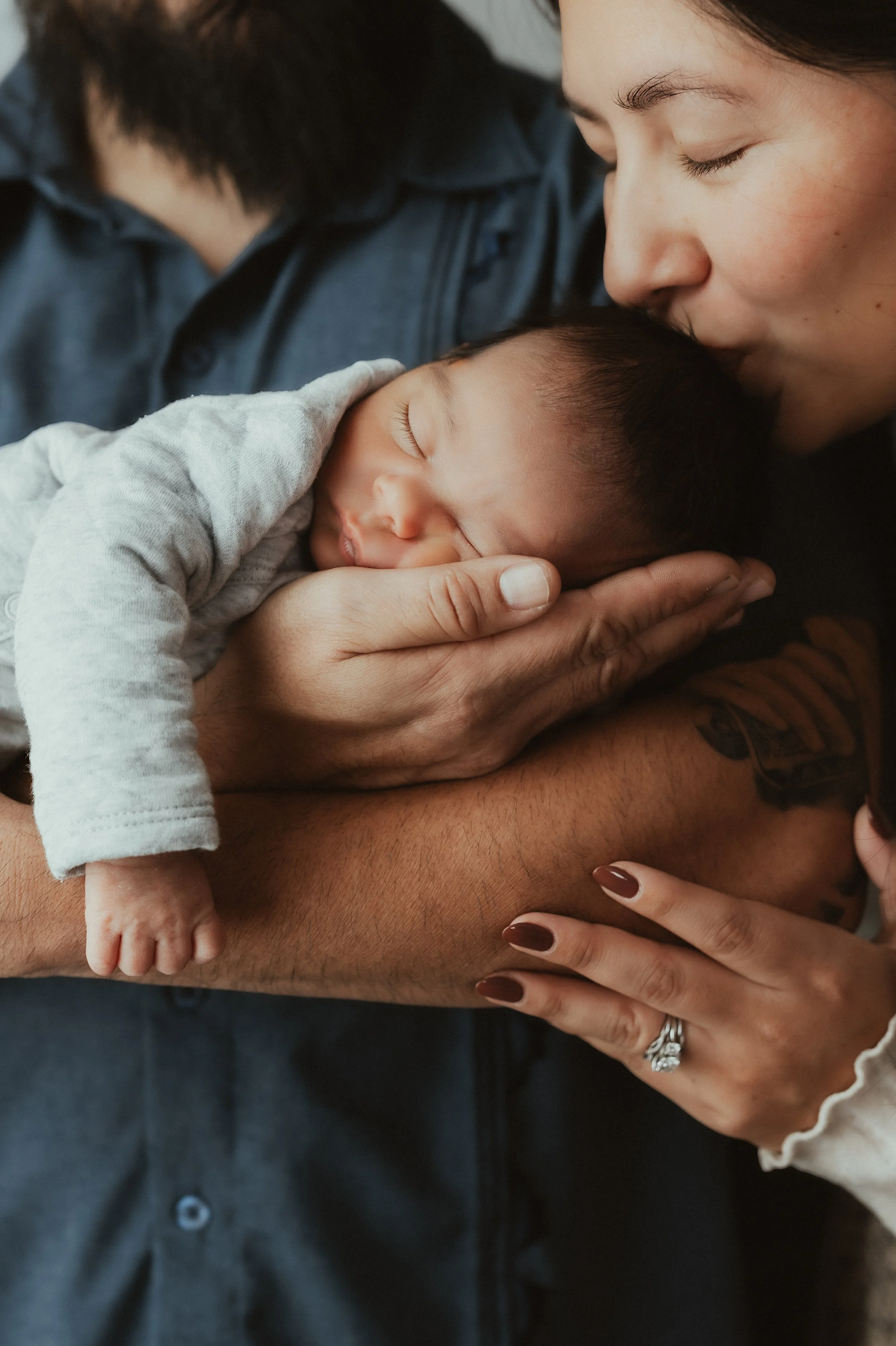 A woman is holding a sleeping baby close to her chest, gently kissing the baby's head. A man is partially visible in the background, wearing a dark shirt. The woman has manicured nails and is wearing a ring. The scene depicts a tender moment of affec