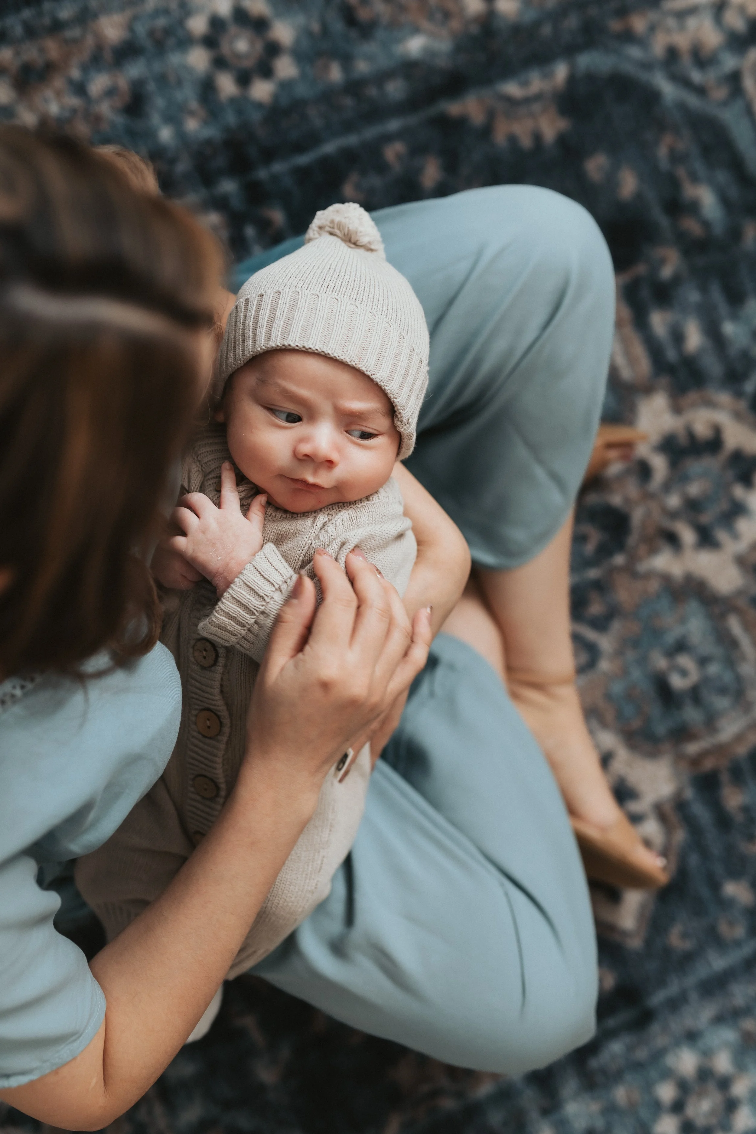 A woman holding a baby while sitting on a patterned rug, with the woman partially visible and the baby wearing a beige hat and sweater, looking to the side.