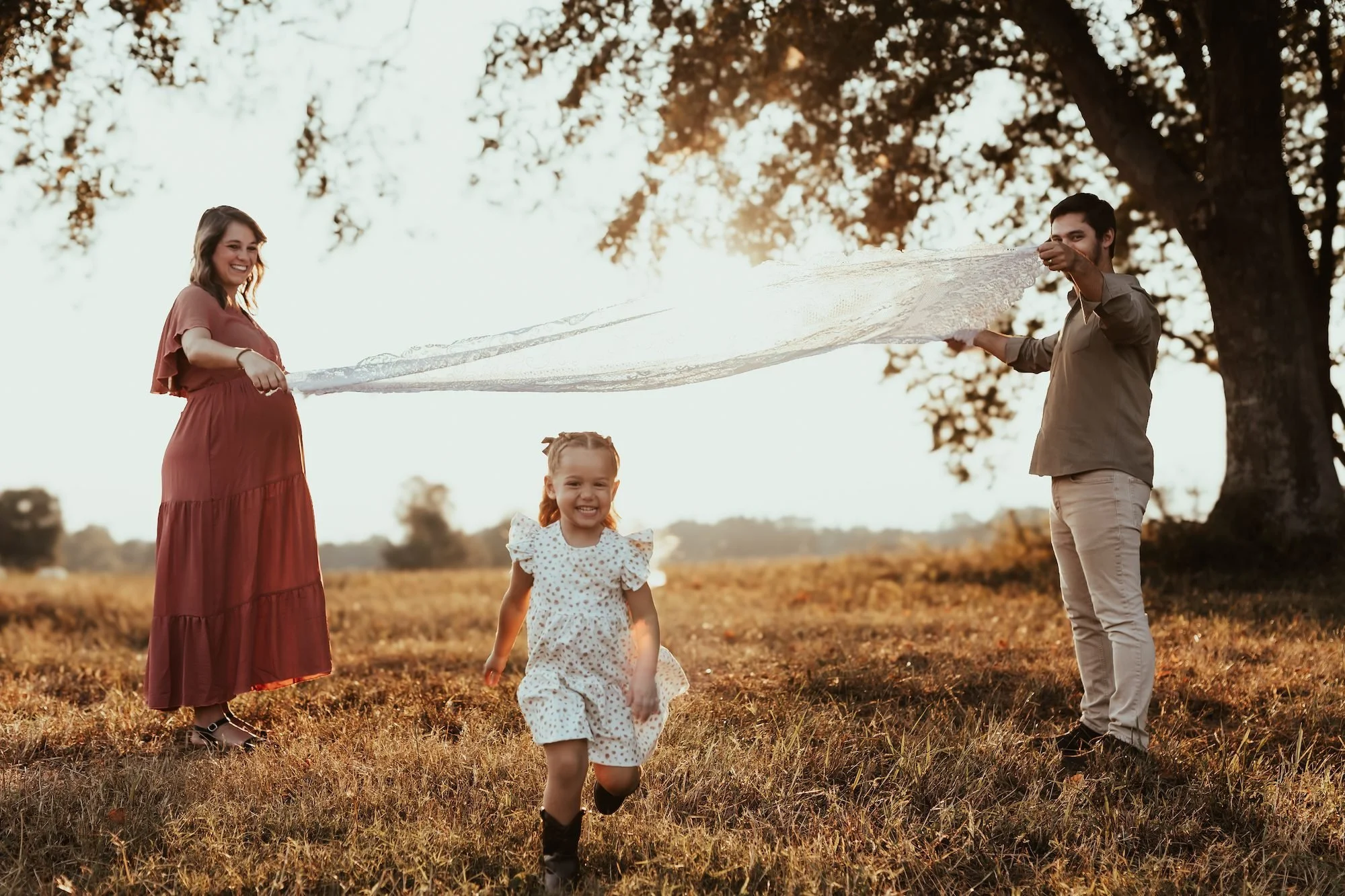 Family playing outdoors in a field during sunset, with a woman holding a lace fabric, a young girl running in front, and a man holding the other end of the fabric.