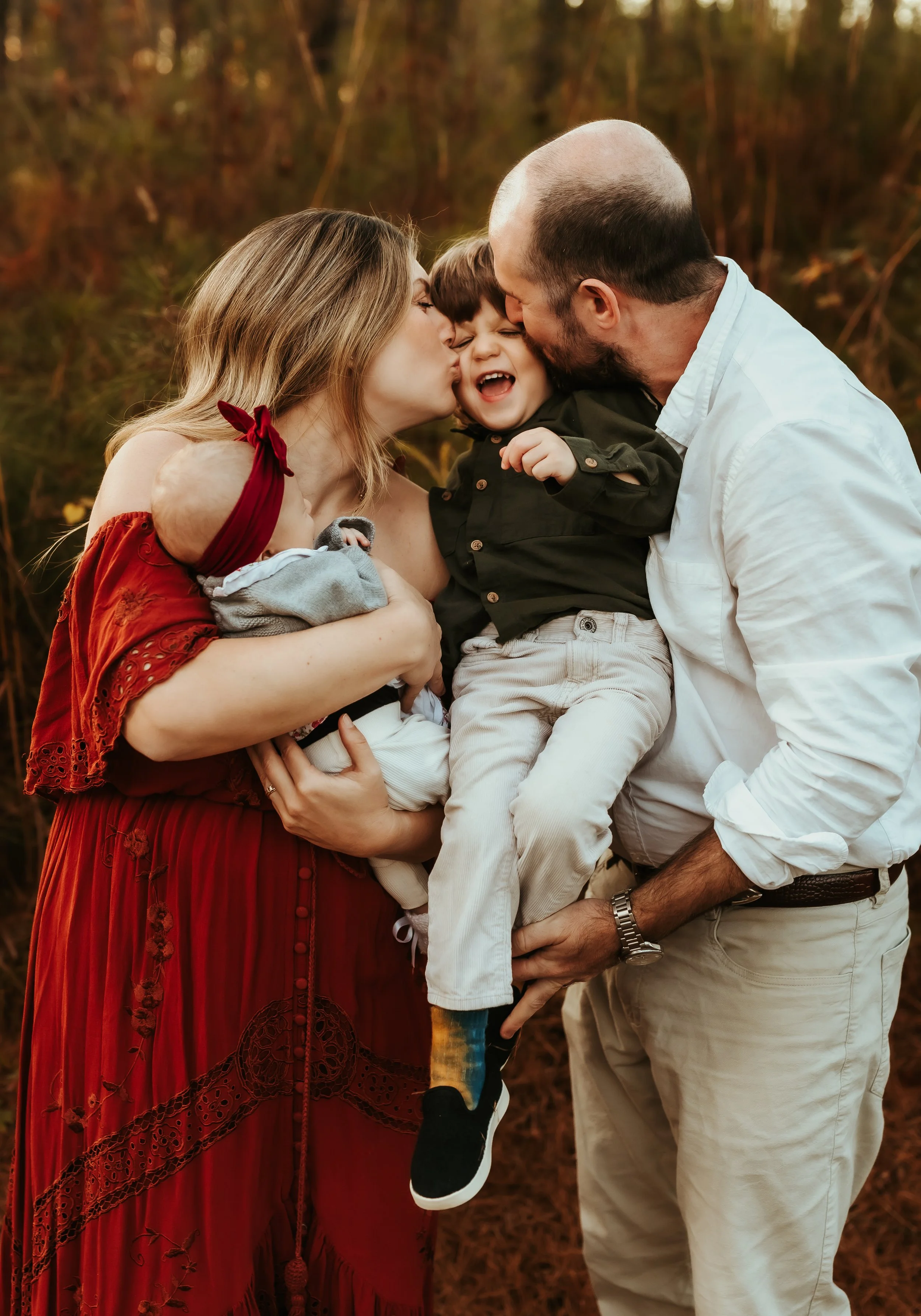 A family of four outside during fall, kissing and hugging each other. The mother is holding a baby while the father is holding an older child. They are all smiling and sharing a loving moment.