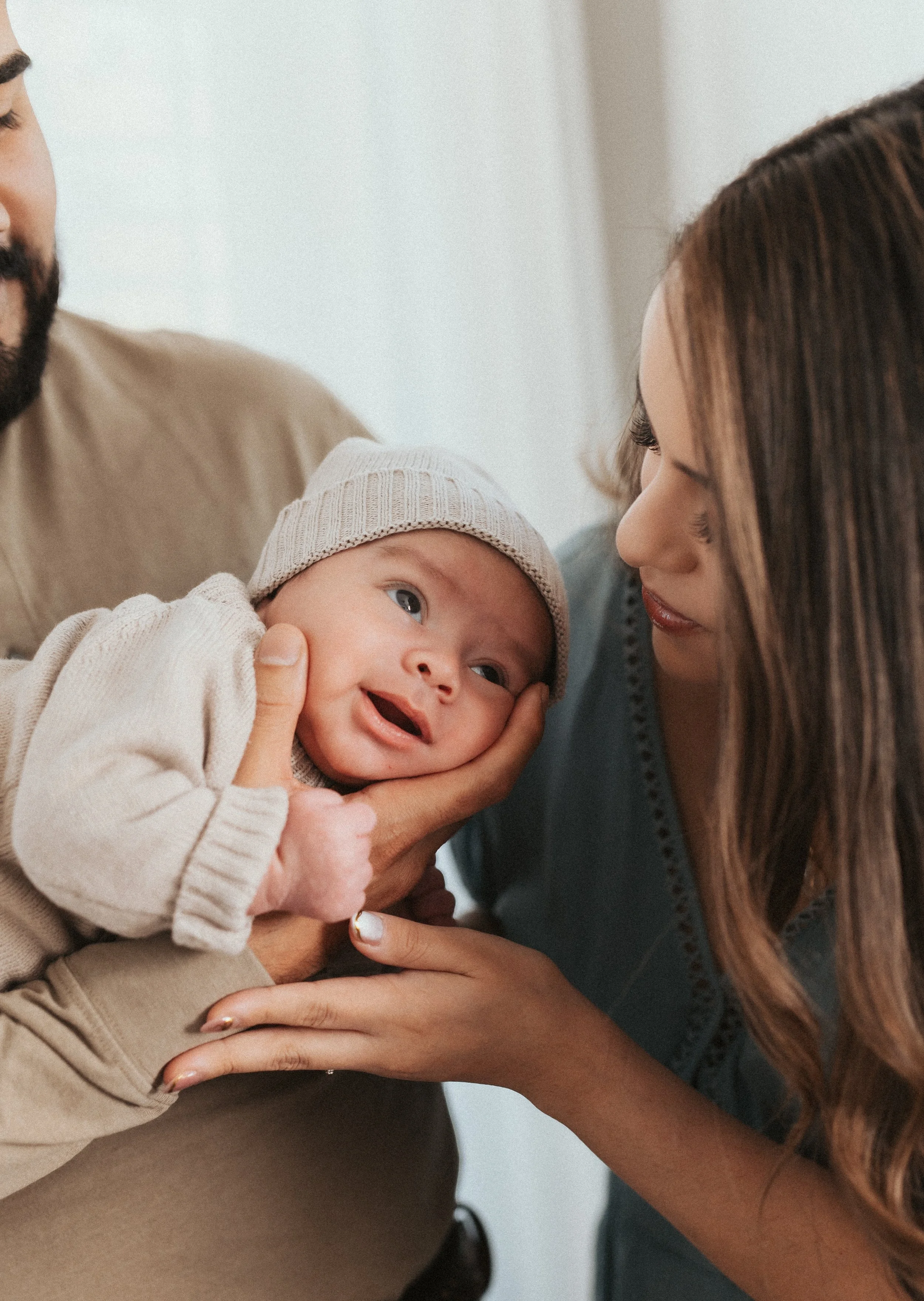 A woman gently touching a smiling baby with a knit hat, with a man partially visible in the background.