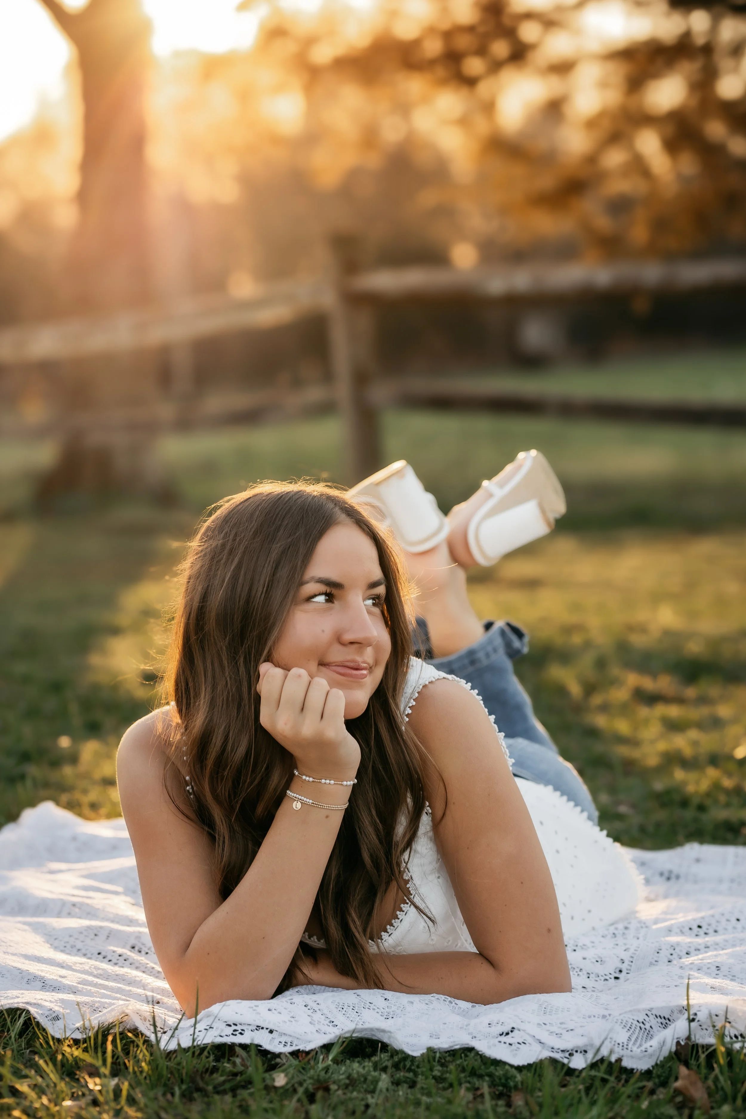 A young woman with long brown hair lying on a white lace blanket on grass, resting her chin on her hand, smiling, outdoors in a park during sunset, with trees in the background.
