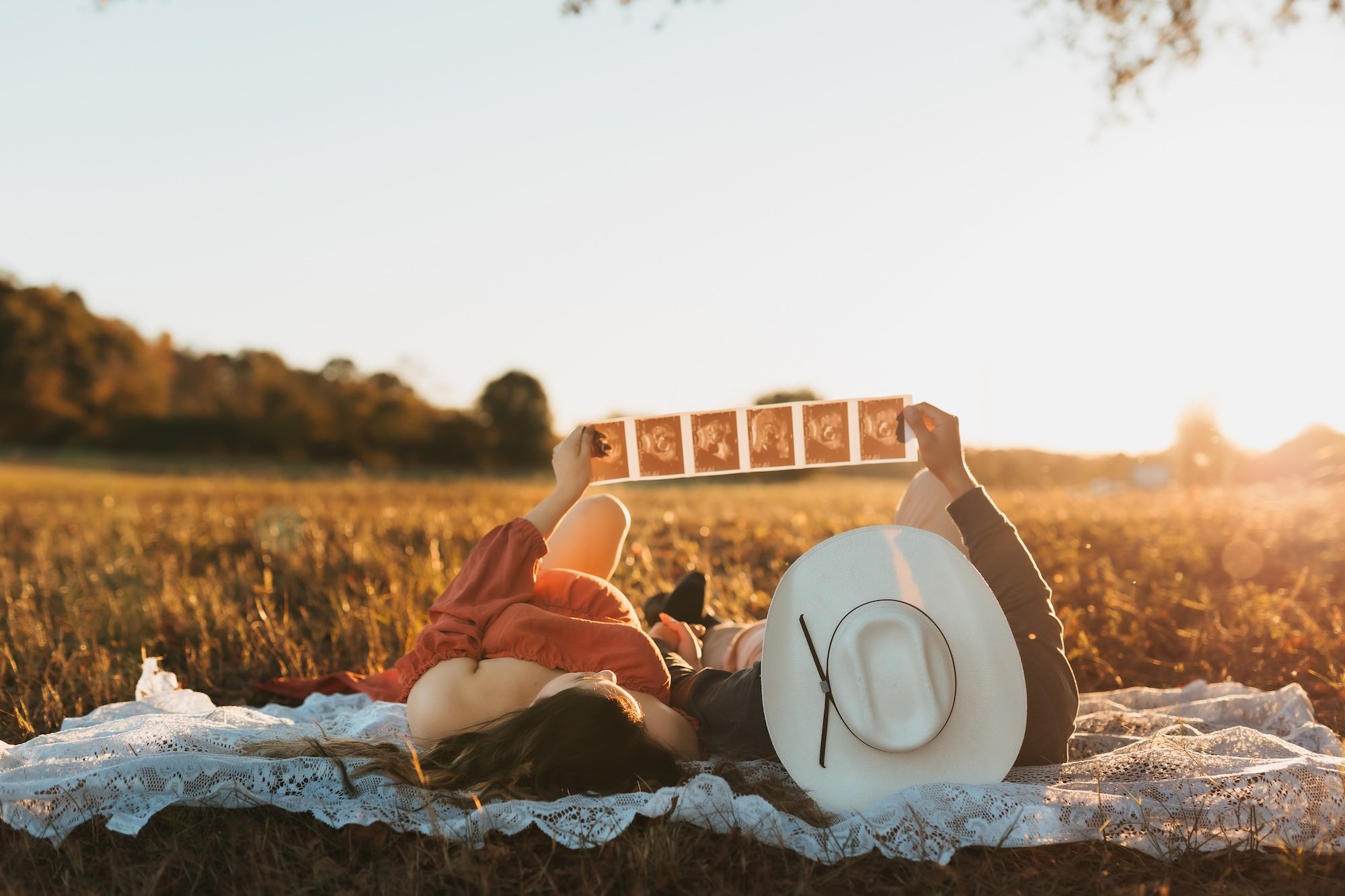 A woman lying on a lace blanket in a field during sunset, holding a strip of film negatives, with a white sun hat nearby.