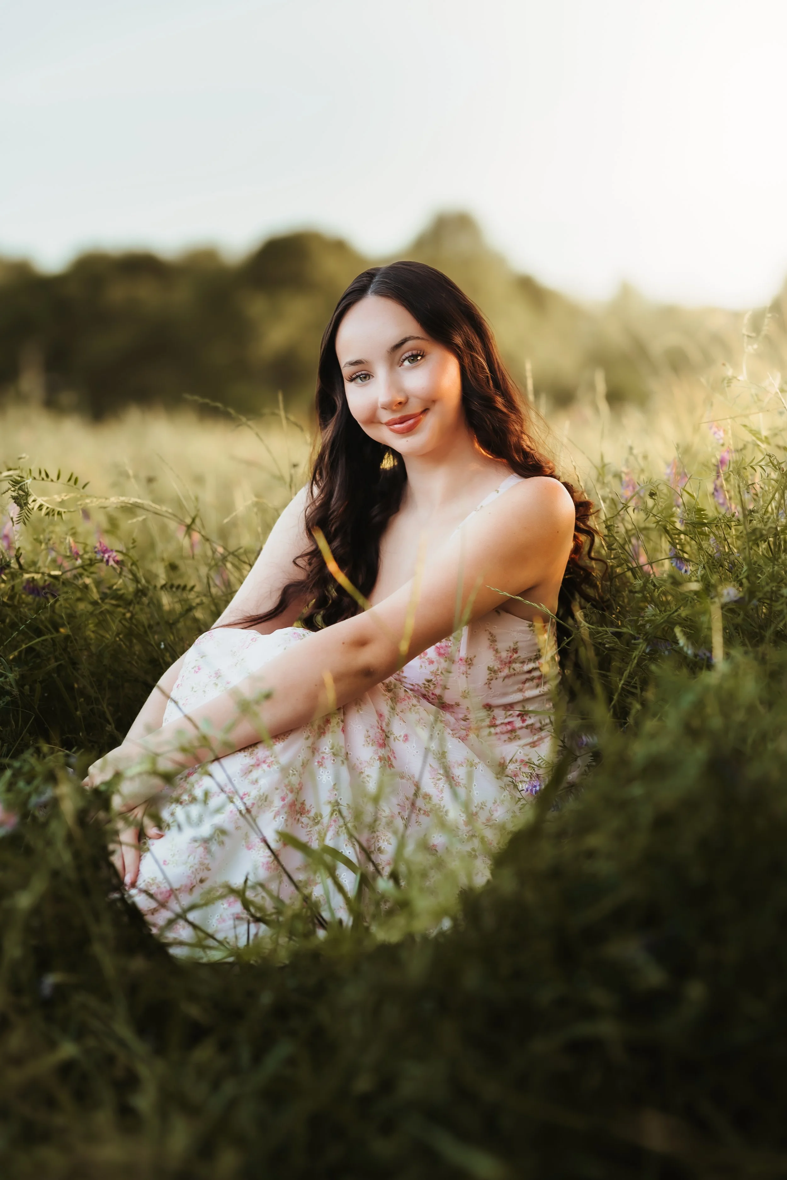 A young woman with long, dark hair, wearing a light-colored floral dress, sitting in a field of tall grass and wildflowers with a blurred background of trees and sky, smiling at the camera during sunset.