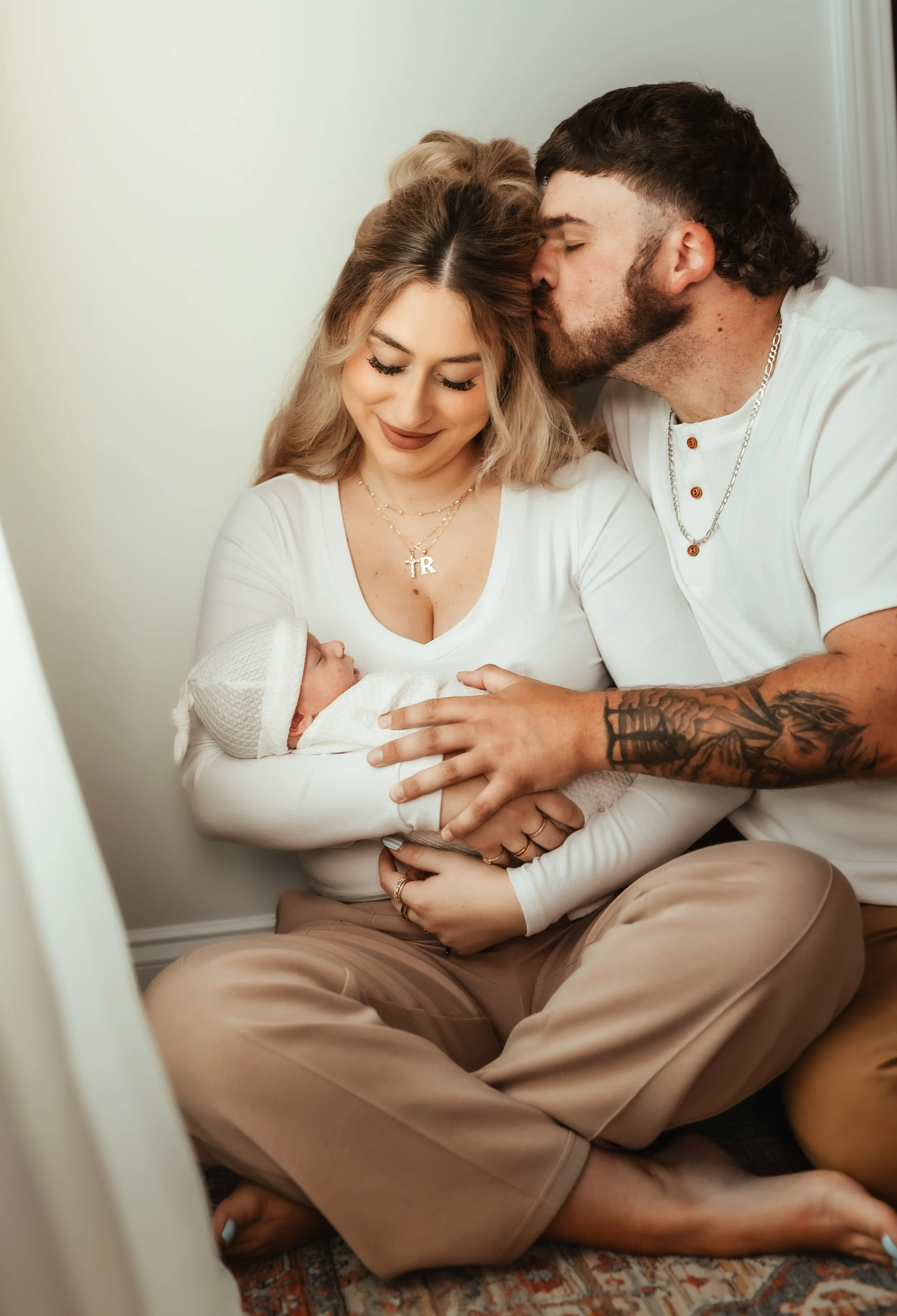 A couple sitting on the floor holding a newborn baby, with the man kissing the woman's forehead.