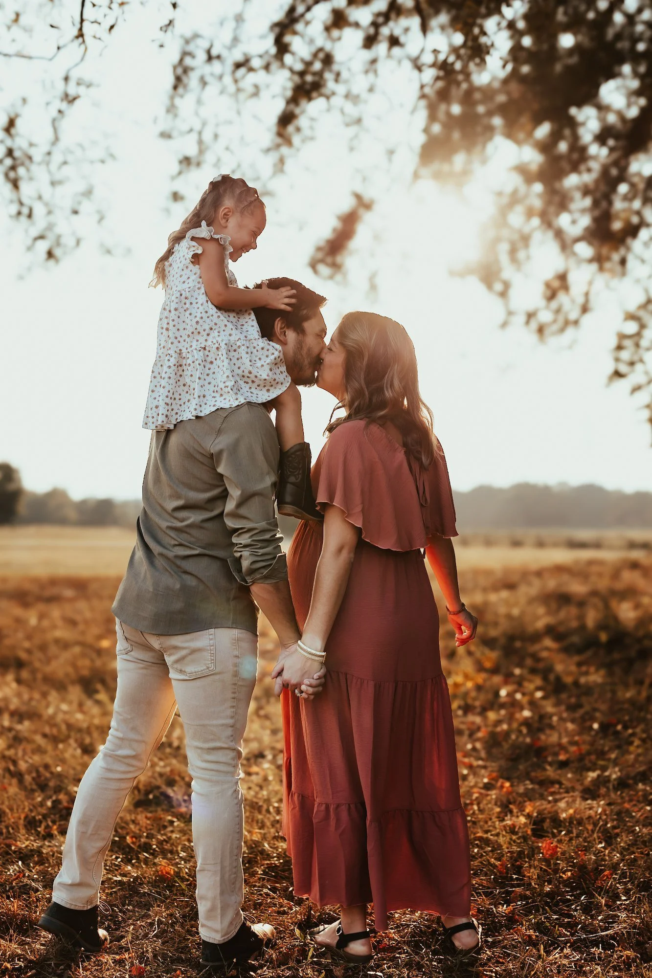 A family of three sharing a kiss outdoors in a field during sunset, with trees in the background.