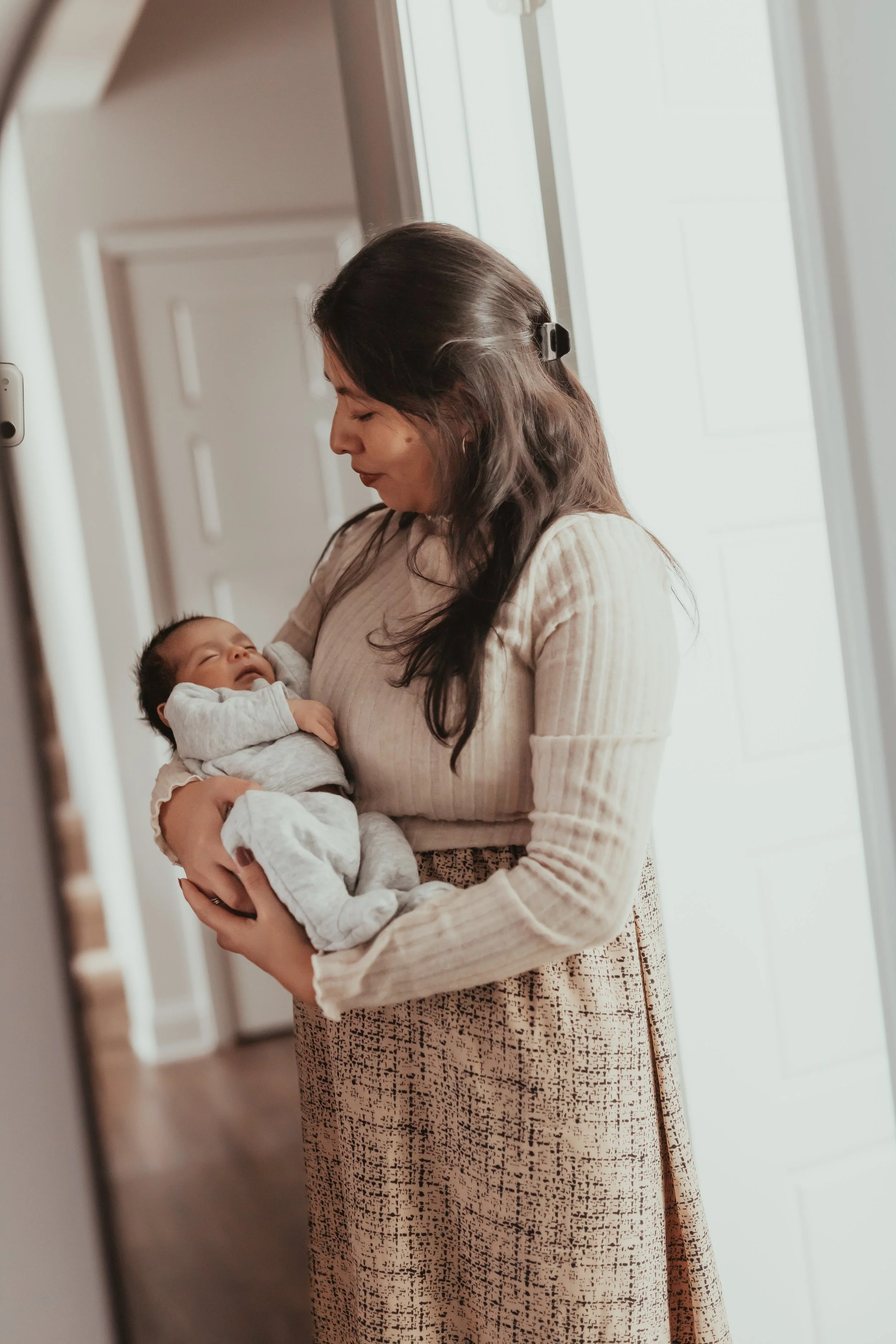 A woman with long dark hair wearing a beige long-sleeve top and a patterned skirt holding a newborn baby in a cozy home setting near a door and window, with soft natural lighting.