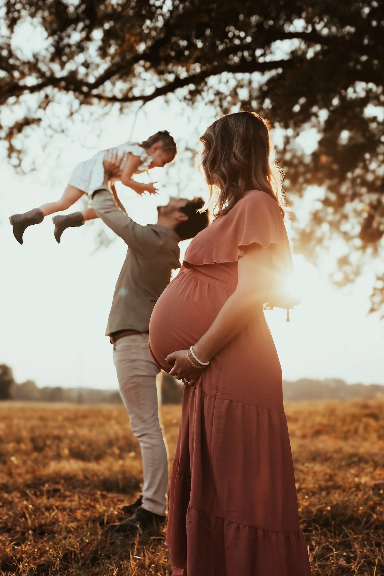 A pregnant woman in a long, flowing dress stands in a field at sunset, holding her belly, while a man behind her lifts a young girl, possibly their daughter, into the air as they all enjoy a moment outdoors.