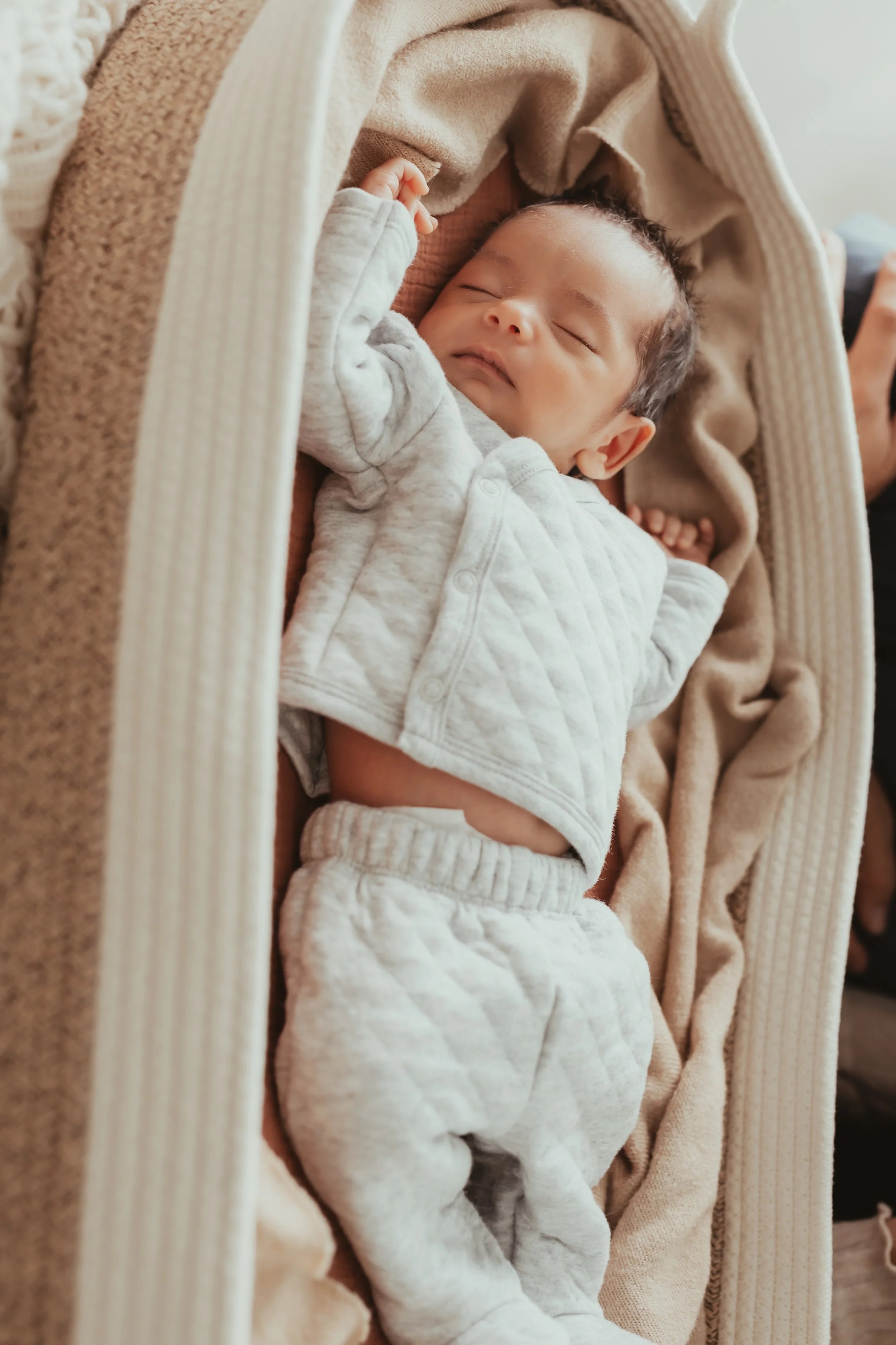 A sleeping baby with curly hair, dressed in a light gray quilted outfit, lying on a soft beige blanket.