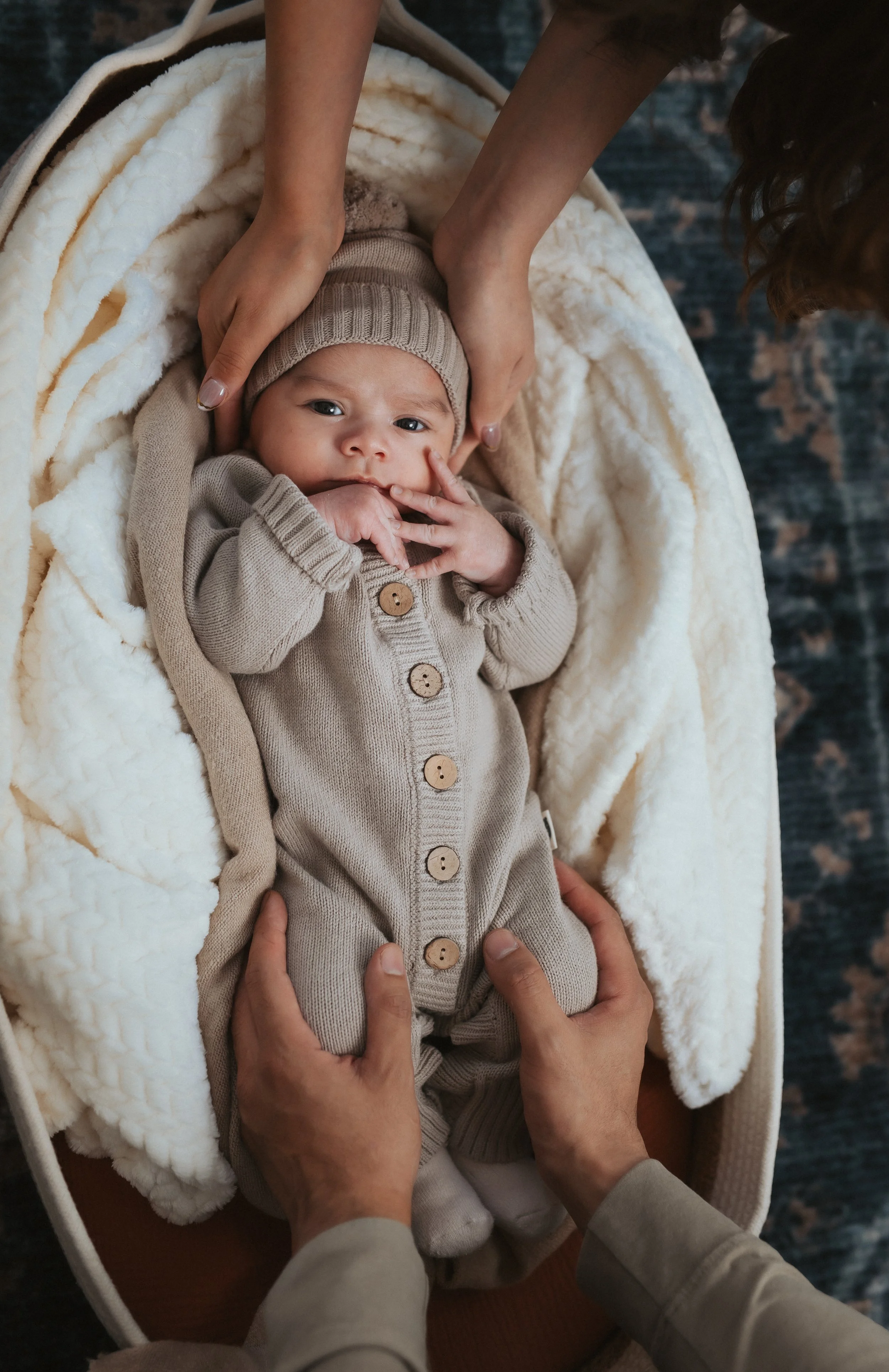 A baby lying on a plush blanket in a bassinet, being gently held and comforted by adult hands. The baby is wearing a beige knitted hat and a matching beige outfit with wooden buttons.