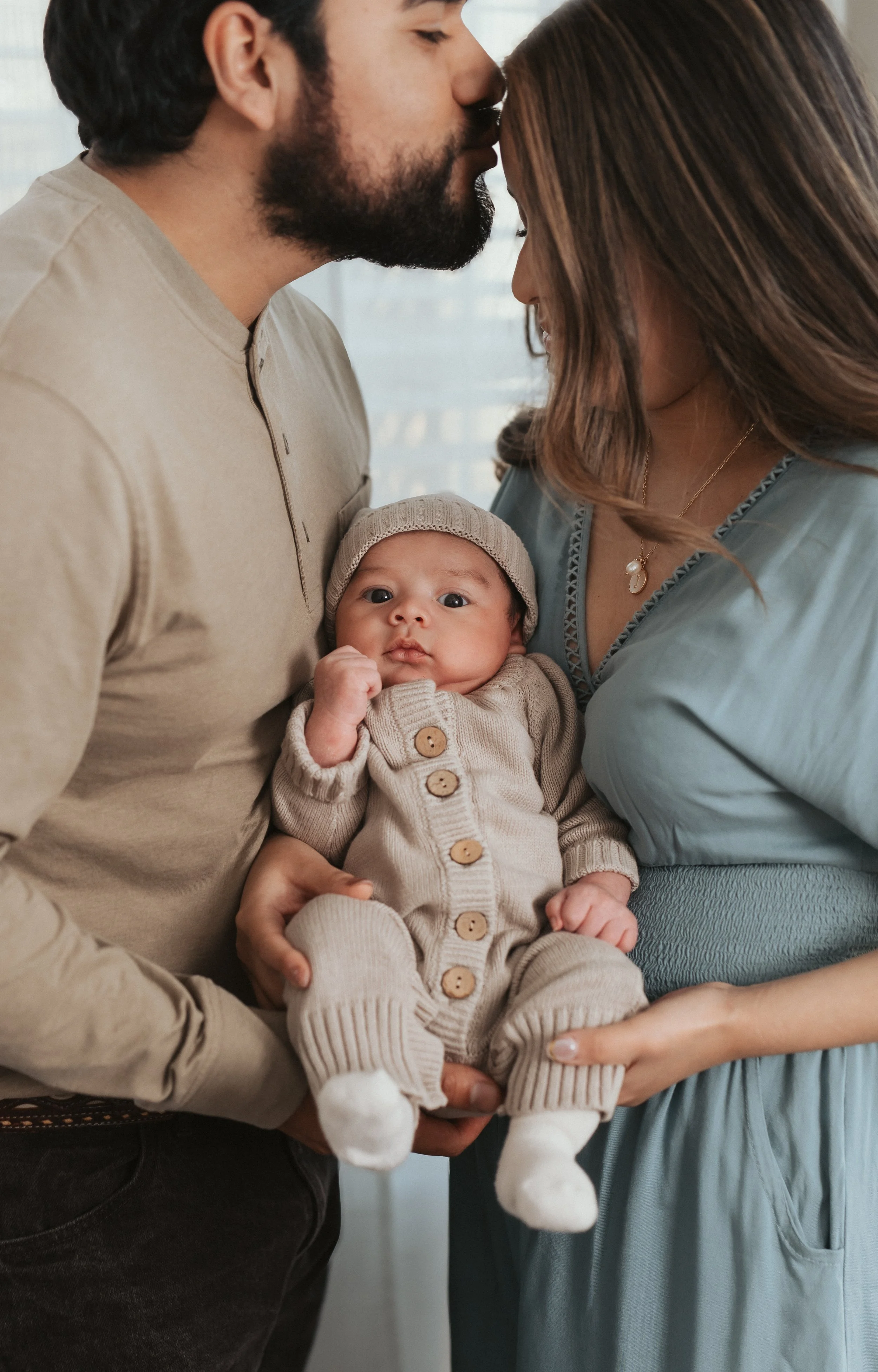 A couple holding a baby, touching foreheads with their eyes closed. The baby is looking at the camera, wearing a beige knit hat and a matching beige outfit with wooden buttons. The couple is dressed in neutral tones, and the background is softly blur