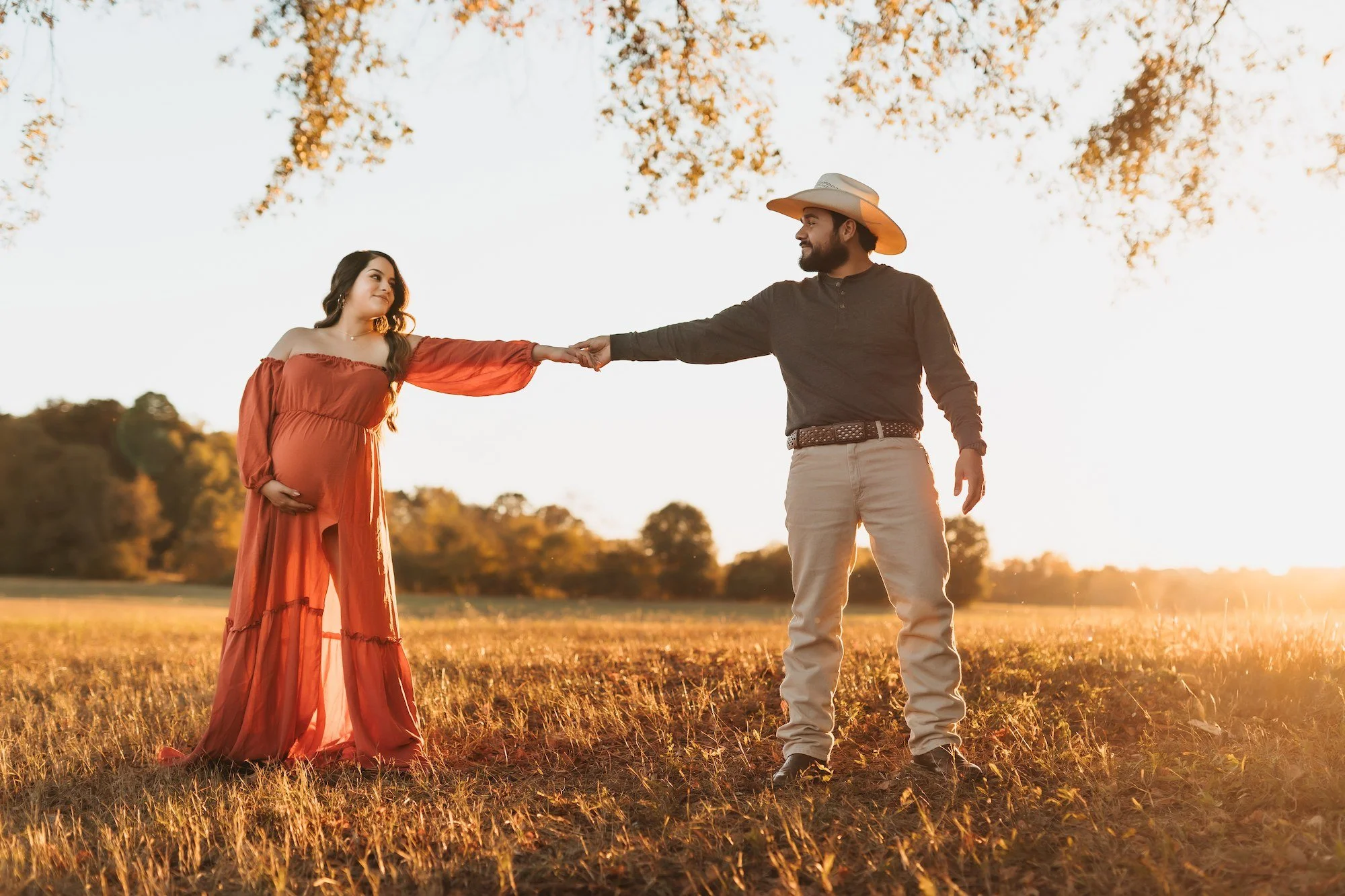 A pregnant woman in an orange dress holding her belly, standing outdoors during sunset, with a man in a cowboy hat reaching out to her with his arm extended.