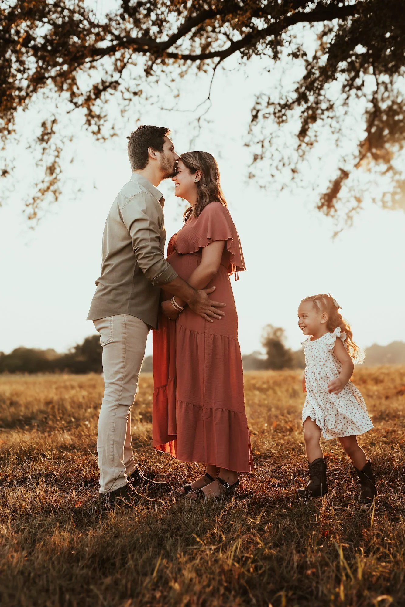 A family of three standing in a field at sunset. The man is kissing the pregnant woman on the forehead, and a little girl is running and smiling nearby.