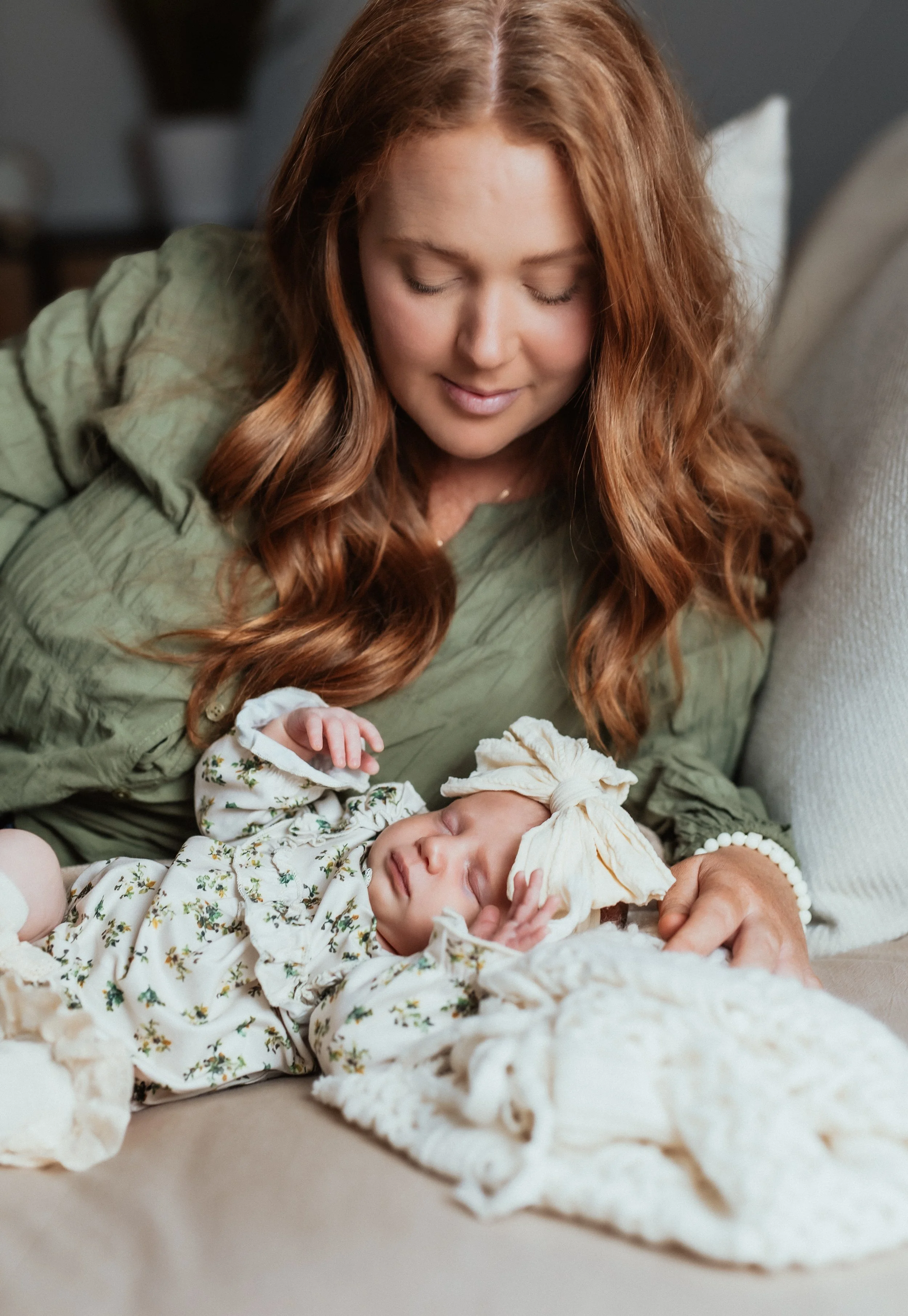 A woman with long red hair and closed eyes is smiling gently while holding a sleeping baby dressed in a light-colored floral onesie and a matching headband, lying on a blanket.