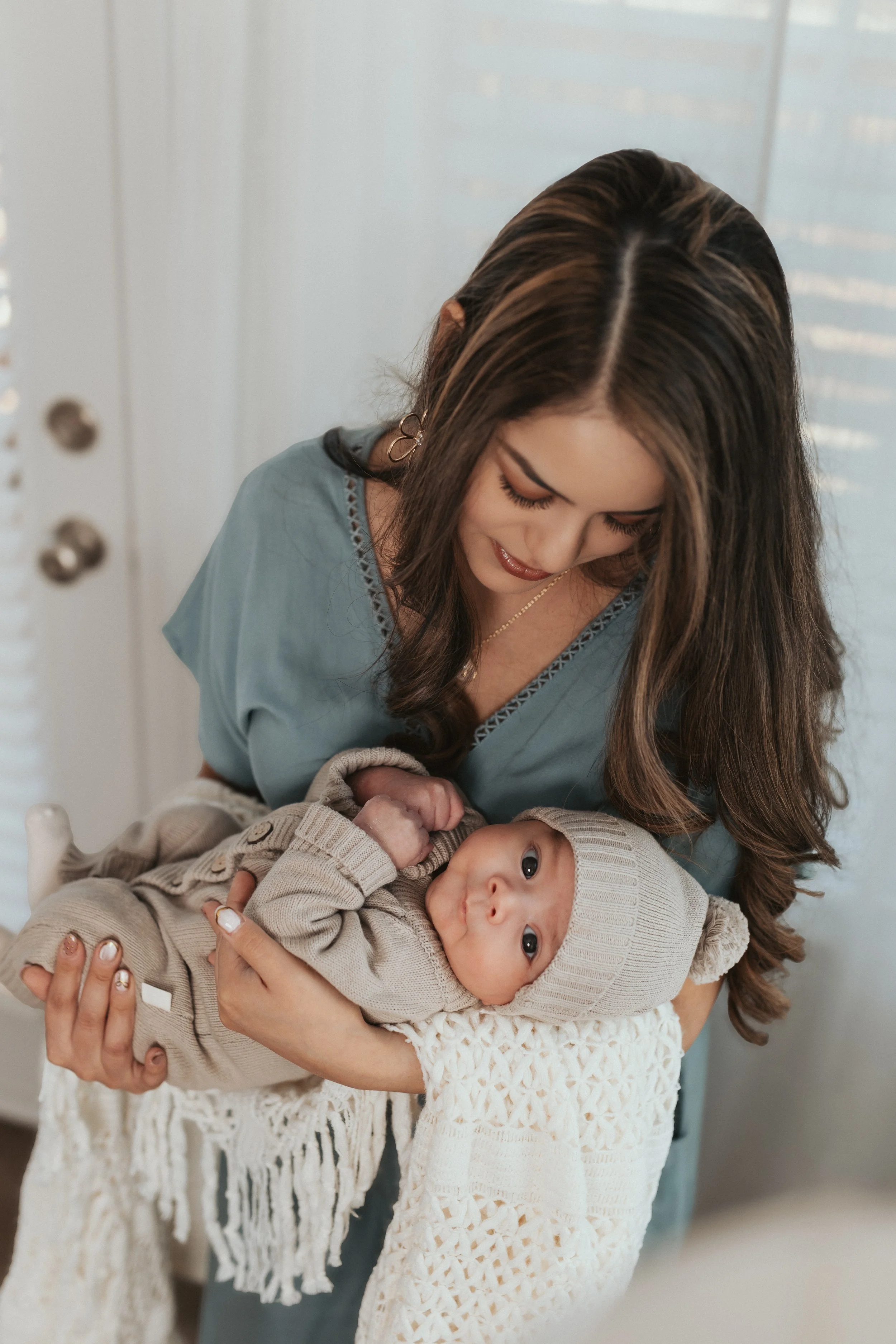 A woman with long brown hair holding a baby with blue eyes, wearing a beige knit hat and cream-colored sweater, in an indoor setting with white walls and a white door.