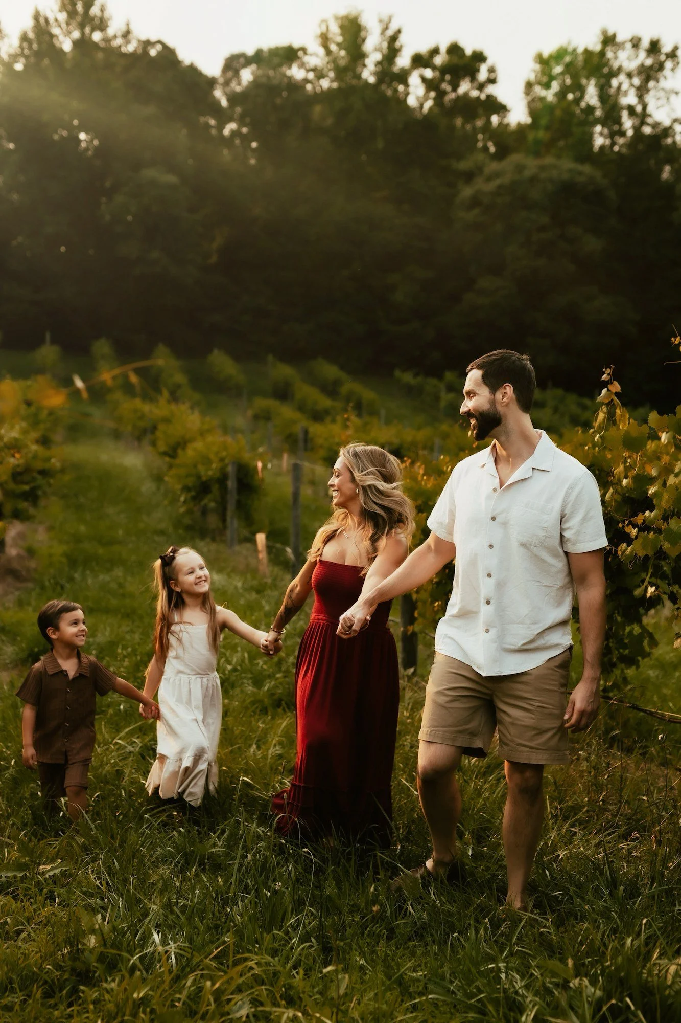 Family of four, including parents and two children, walking through a vineyard at sunset, holding hands and smiling.