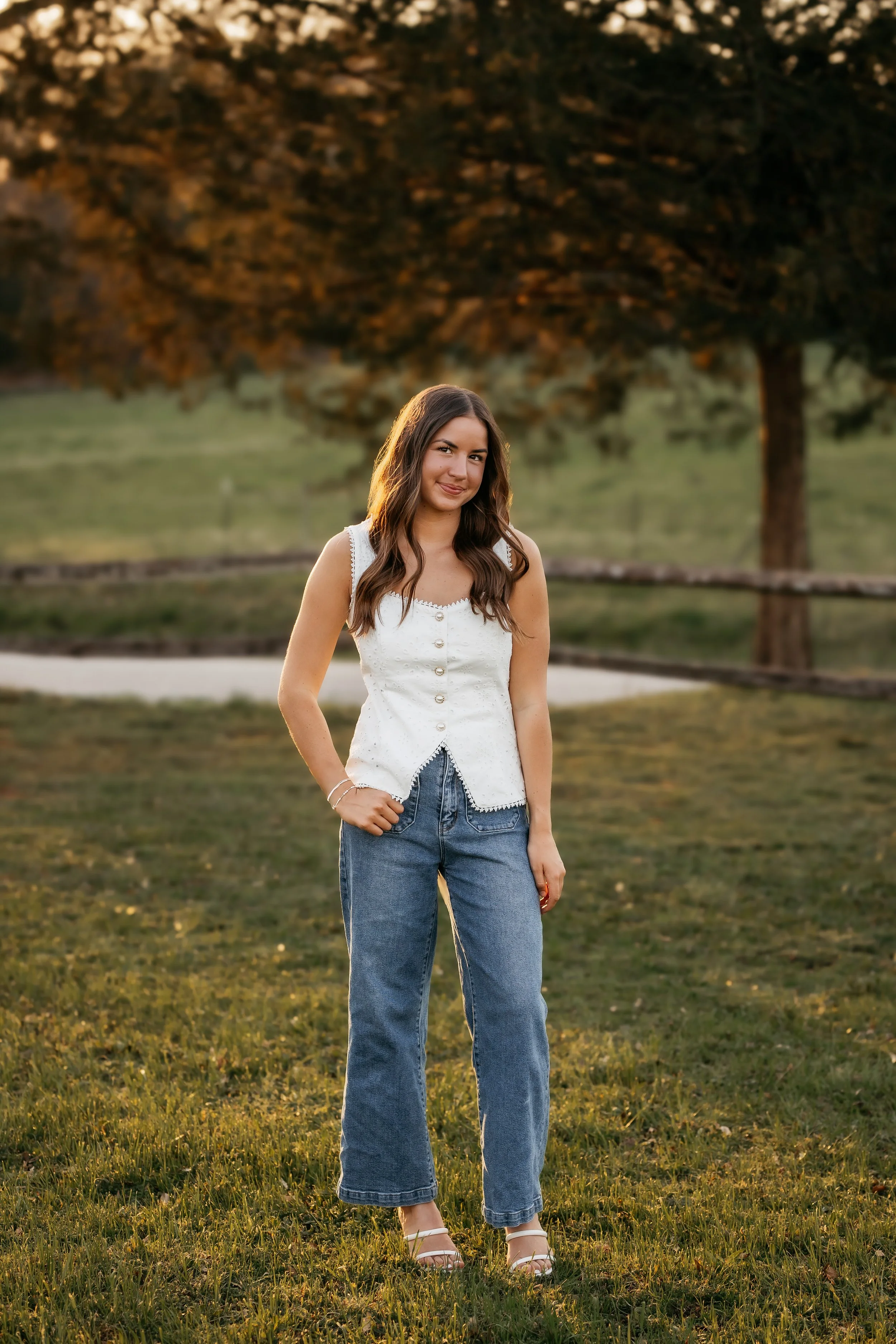 A young woman with long brown hair smiling outdoors during sunset, wearing a white sleeveless top and blue jeans, standing on grass with trees in the background.