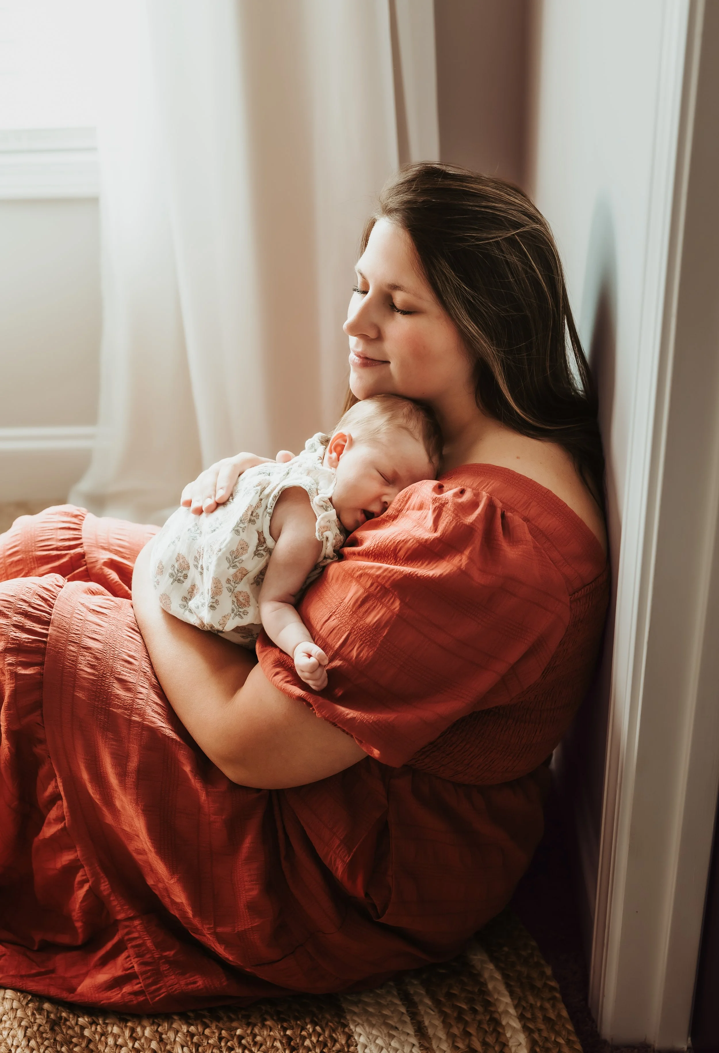 A woman with long dark hair and a reddish-orange dress sits on a rug, holding a sleeping baby with a lace and floral outfit, against a corner of a room with a white wall and curtains.