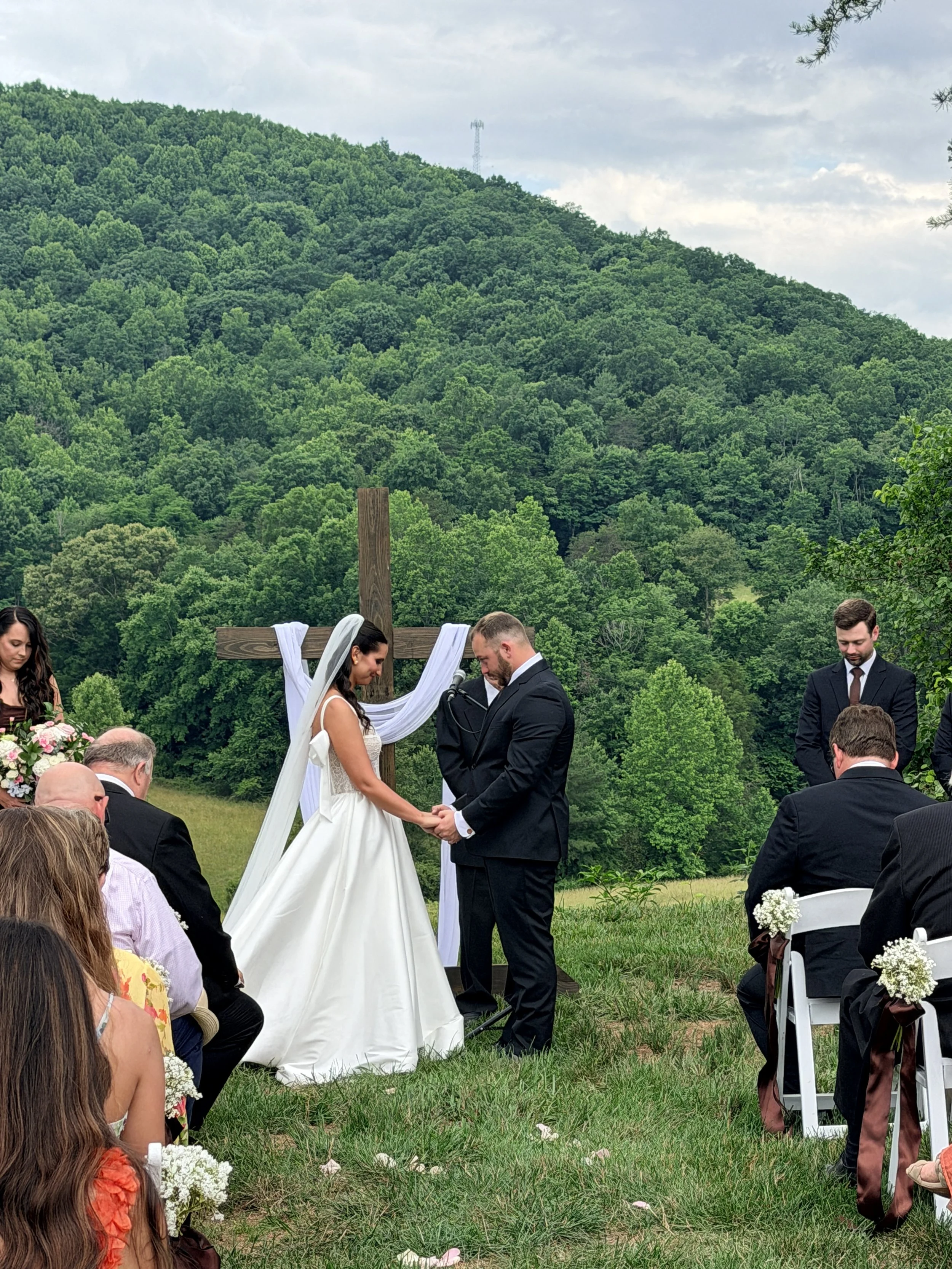 A couple is getting married at mountain view wedding ceremony standing in front of a wooden cross decorated with white fabric, with a lush green mountain and cloudy sky in the background. Guests are seated on either side, watching the ceremony.
