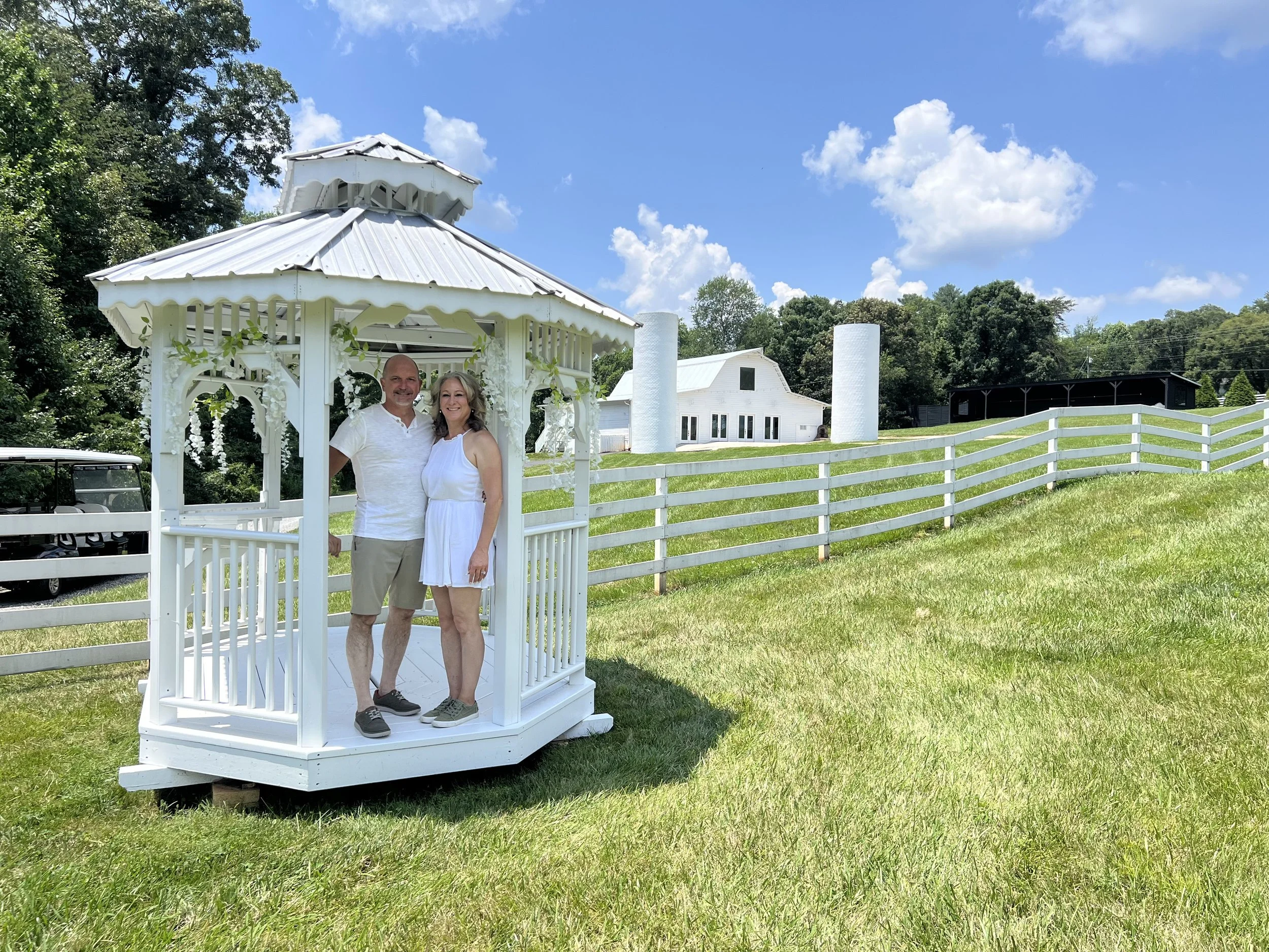 Owners of Redbird Ridge Estate standing in white gazebo with white venue and silos in distance and lush green grass all around them