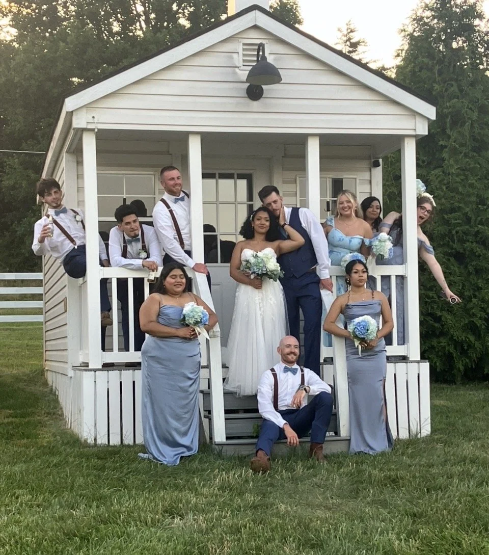 A group of people is gathered on the front porch of a small white house, celebrating a wedding. The bride and groom are in the center, with the bride holding a bouquet of white flowers. The group is dressed in formal attire, with women in matching light blue dresses and men in suspenders and bow ties. Some members of the group are sitting or leaning on the porch railing, while others stand or sit on the front steps. The setting appears to be outdoors, with trees and grass visible in the background at sunset.