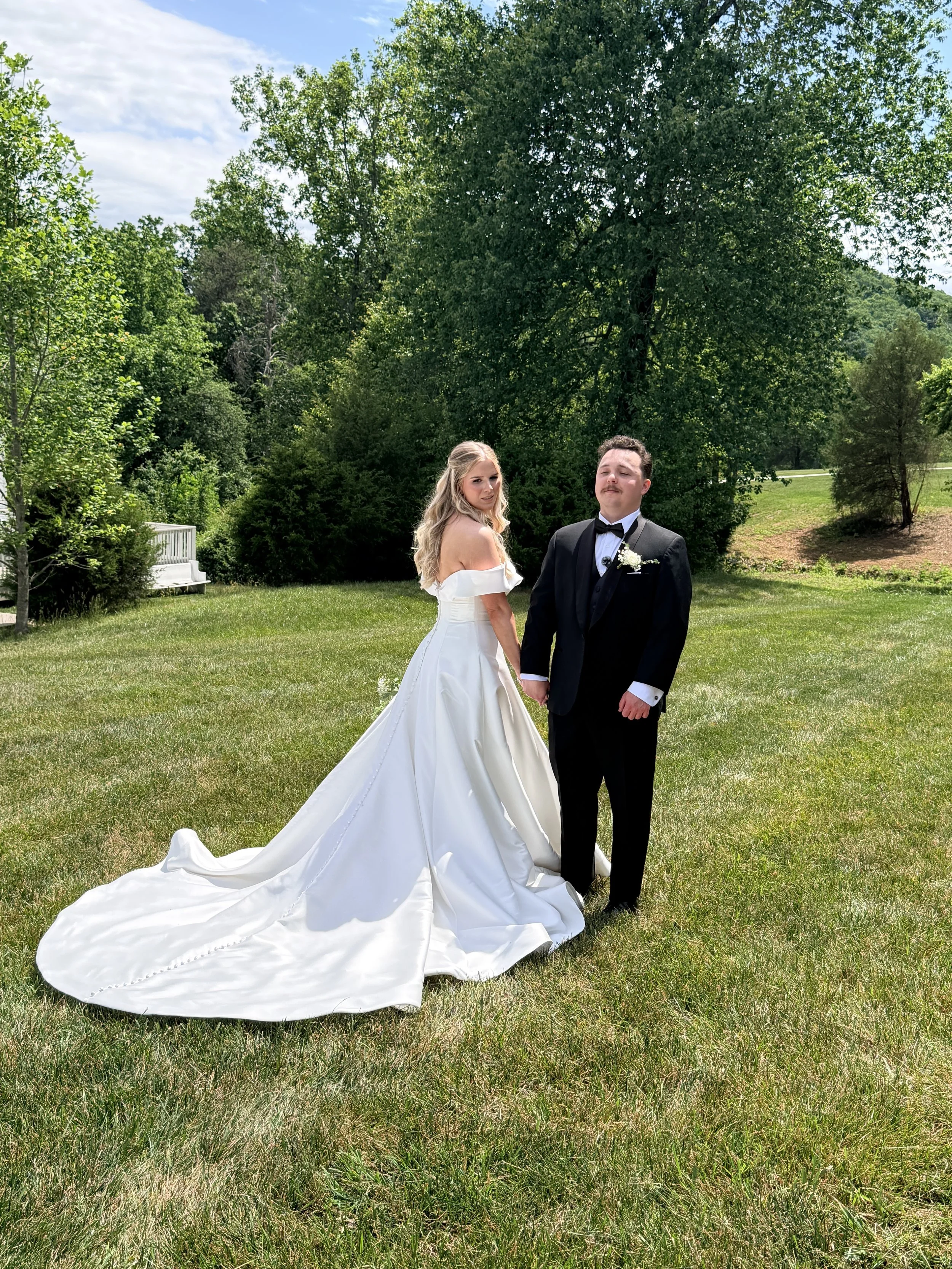 A bride and groom standing hand in hand outdoors in a grassy area with green trees and a partly cloudy sky in the background.