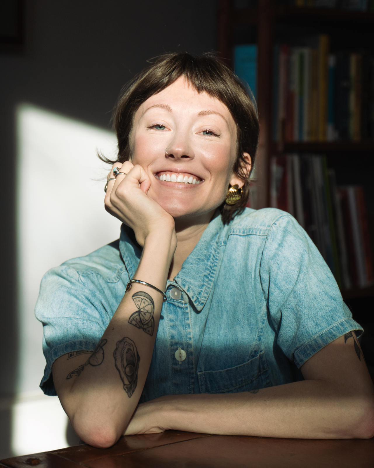A smiling woman with short brown hair, wearing a light denim shirt, gold hoop earrings, and multiple rings. She is sitting indoors with sunlight on her face, in front of a bookshelf.