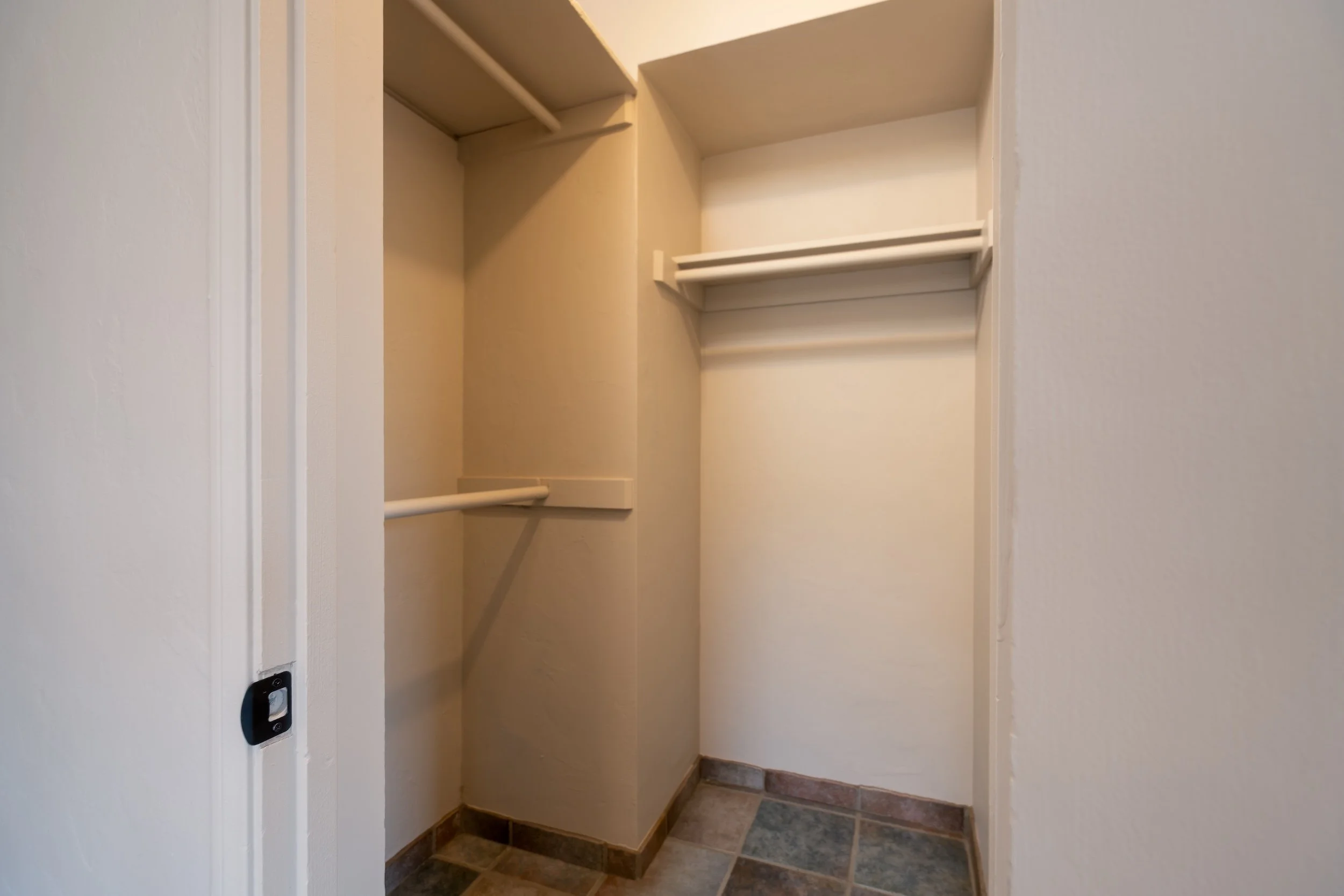Empty closet with beige walls, shelves, and a hanging rod, located in a small hallway with tiled floor.