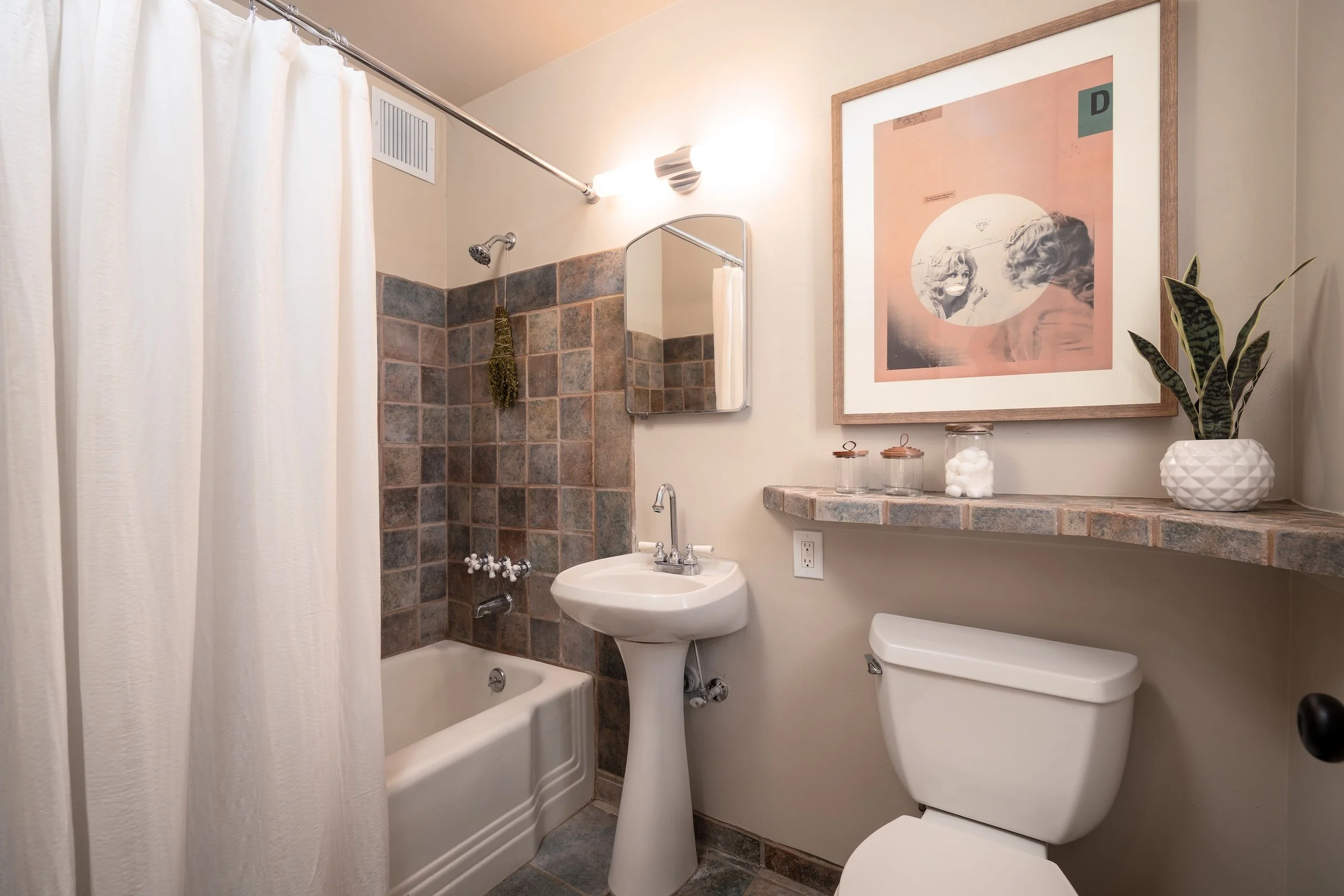 A small bathroom with a bathtub and shower, white shower curtain, tile surround, a small mirror, a sink, a toilet, a framed artwork on the wall, and a potted plant on a counter.