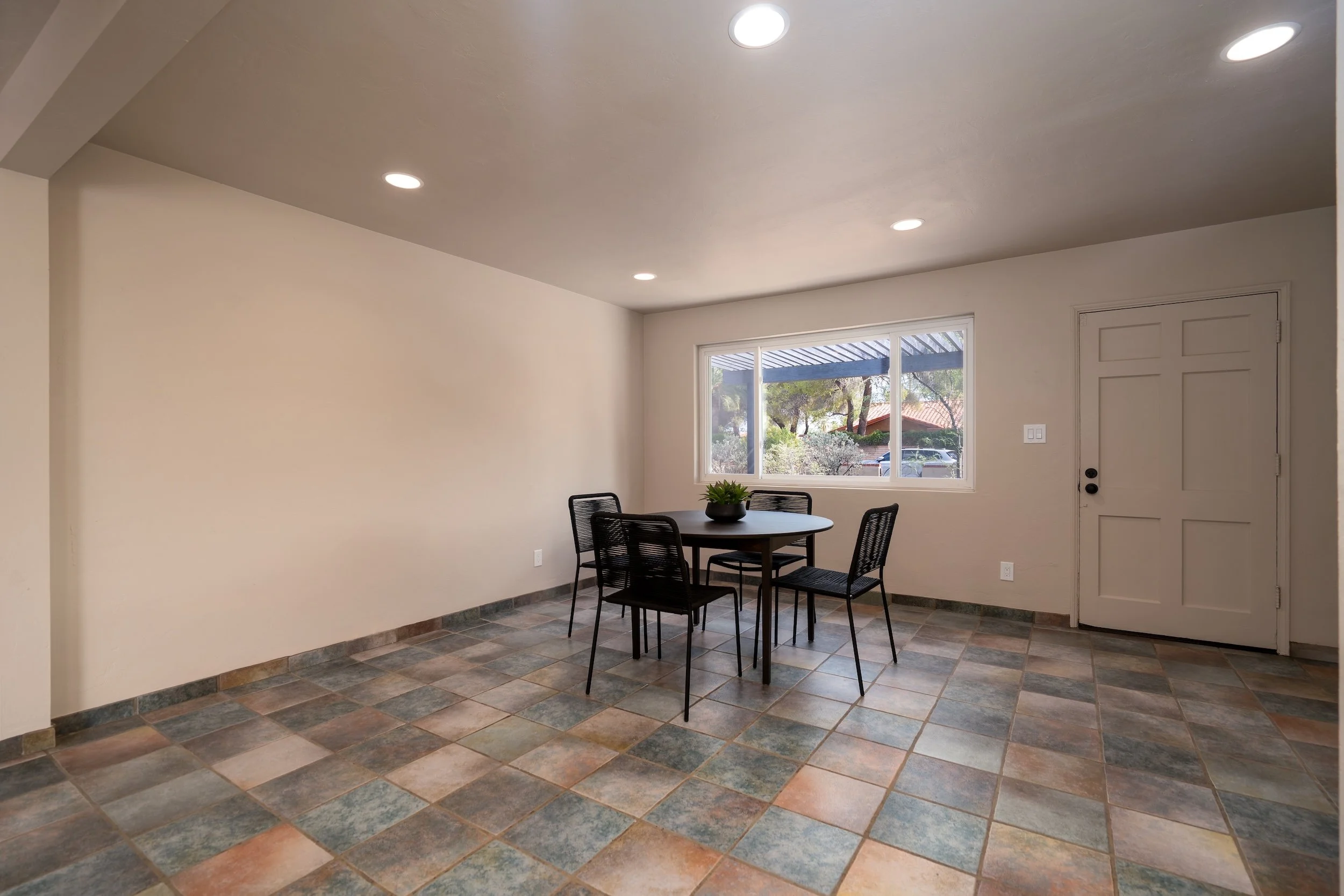 Interior of a room with a small dining table and four black chairs, a window showing trees and neighboring houses, a door, and tiled flooring, with four ceiling lights.